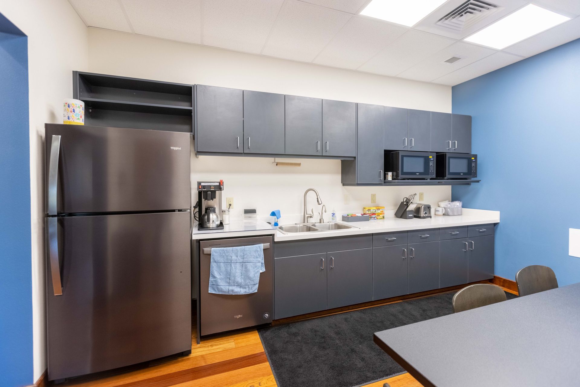 Small office kitchenette with stainless fridge, gray cabinets, sink, microwave, and a table in the foreground