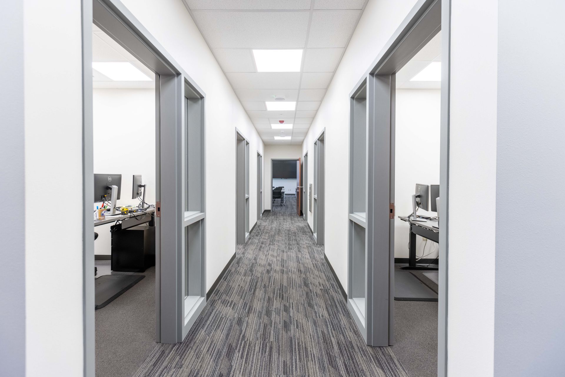 Modern office hallway with open doorways, gray carpet, and bright overhead lights
