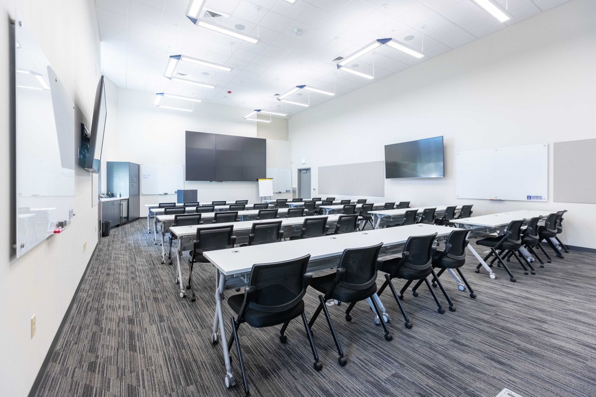 Empty modern classroom with rows of desks, black chairs, large wall screens, and carpeted floor