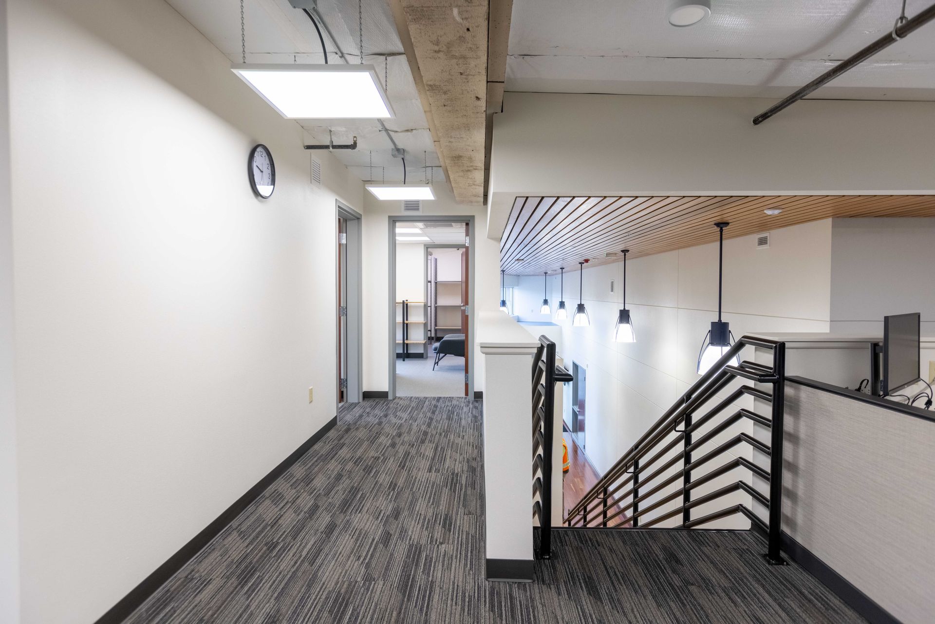 Modern office hallway with gray carpet, white walls, and a stairwell railing overlooking the lower floor