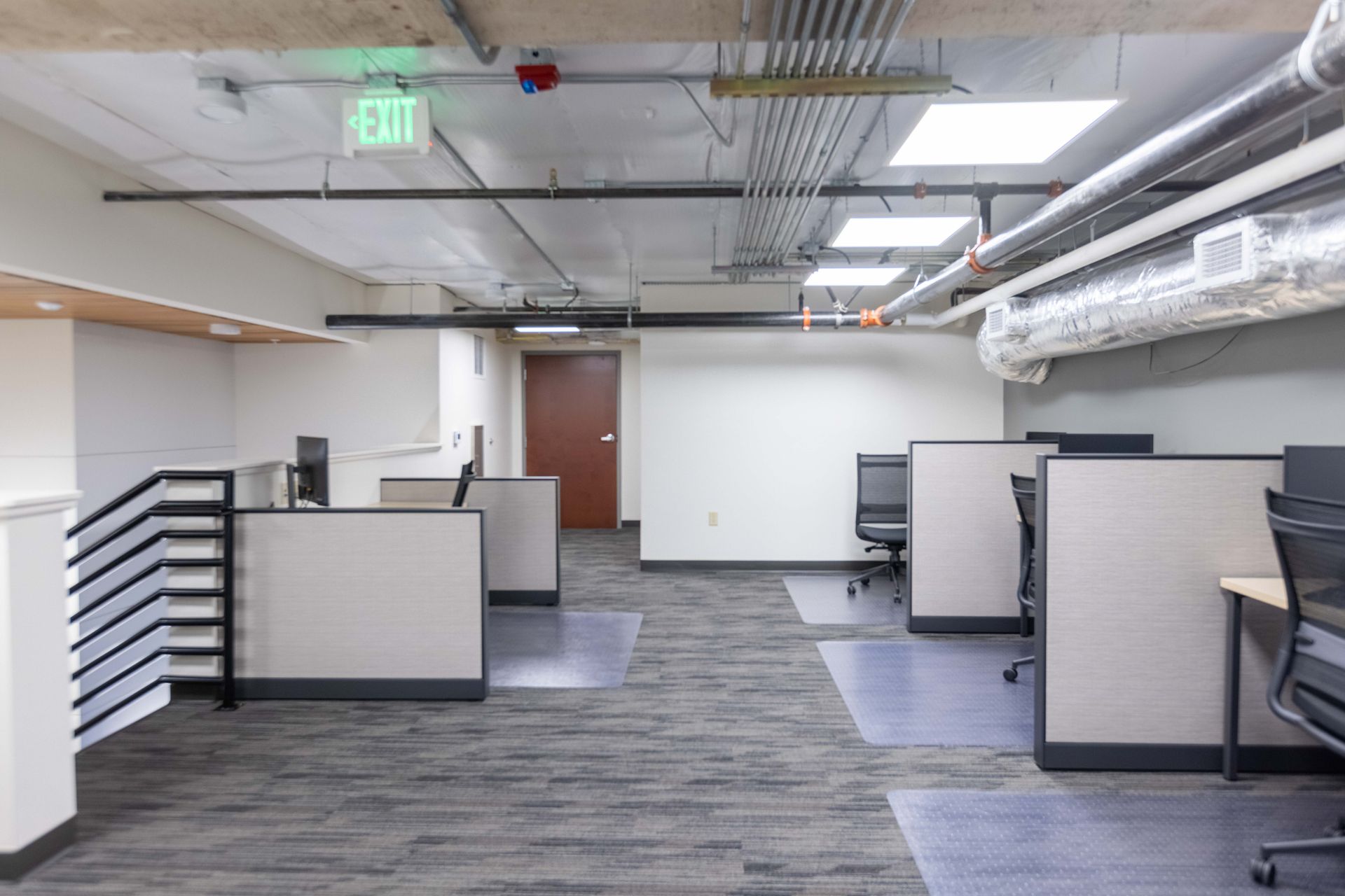 Empty modern office with gray cubicles, carpeted floor, and exposed ceiling lights.