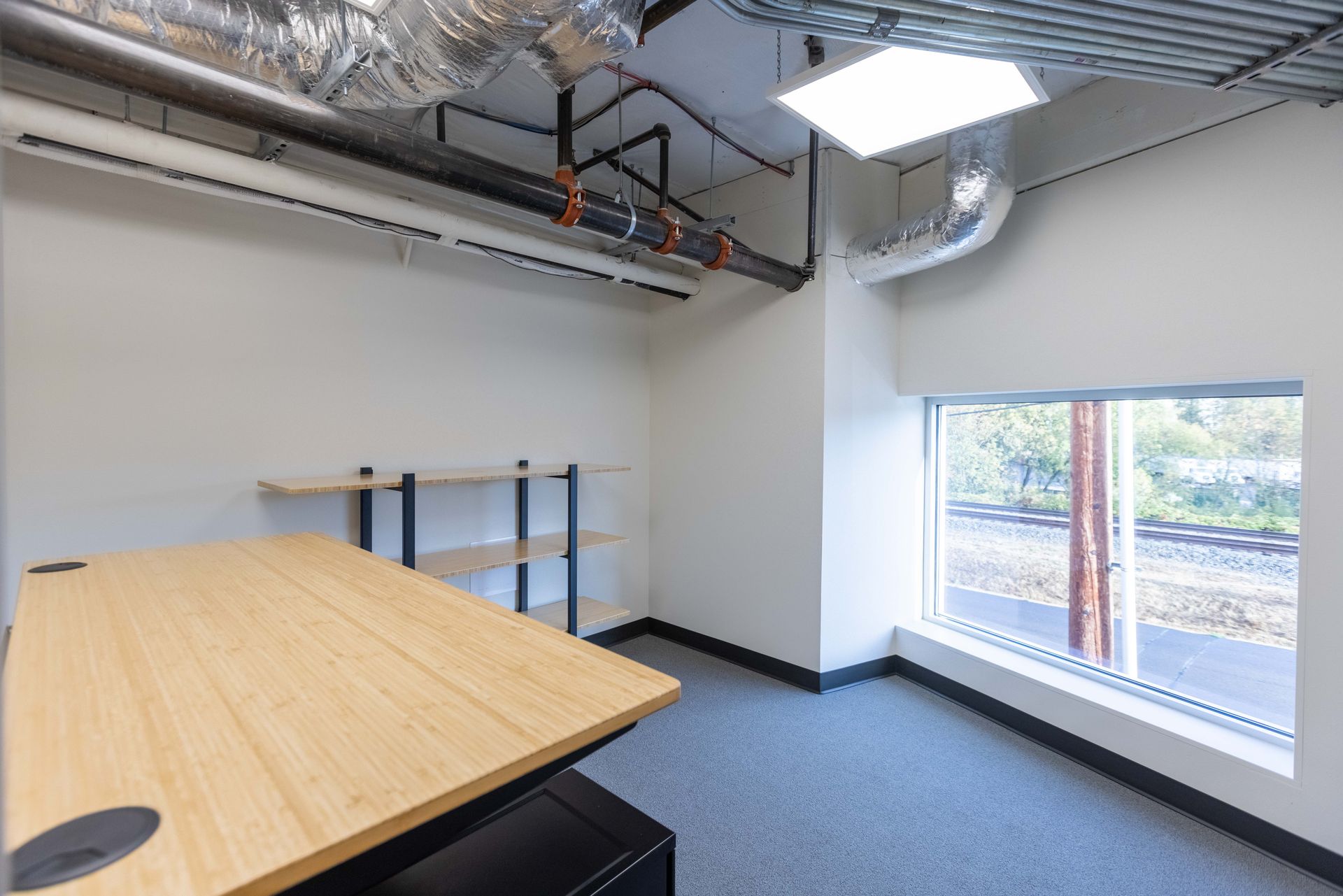 Empty office corner with wood table, metal shelving, exposed ceiling ducts, and a large window with blinds