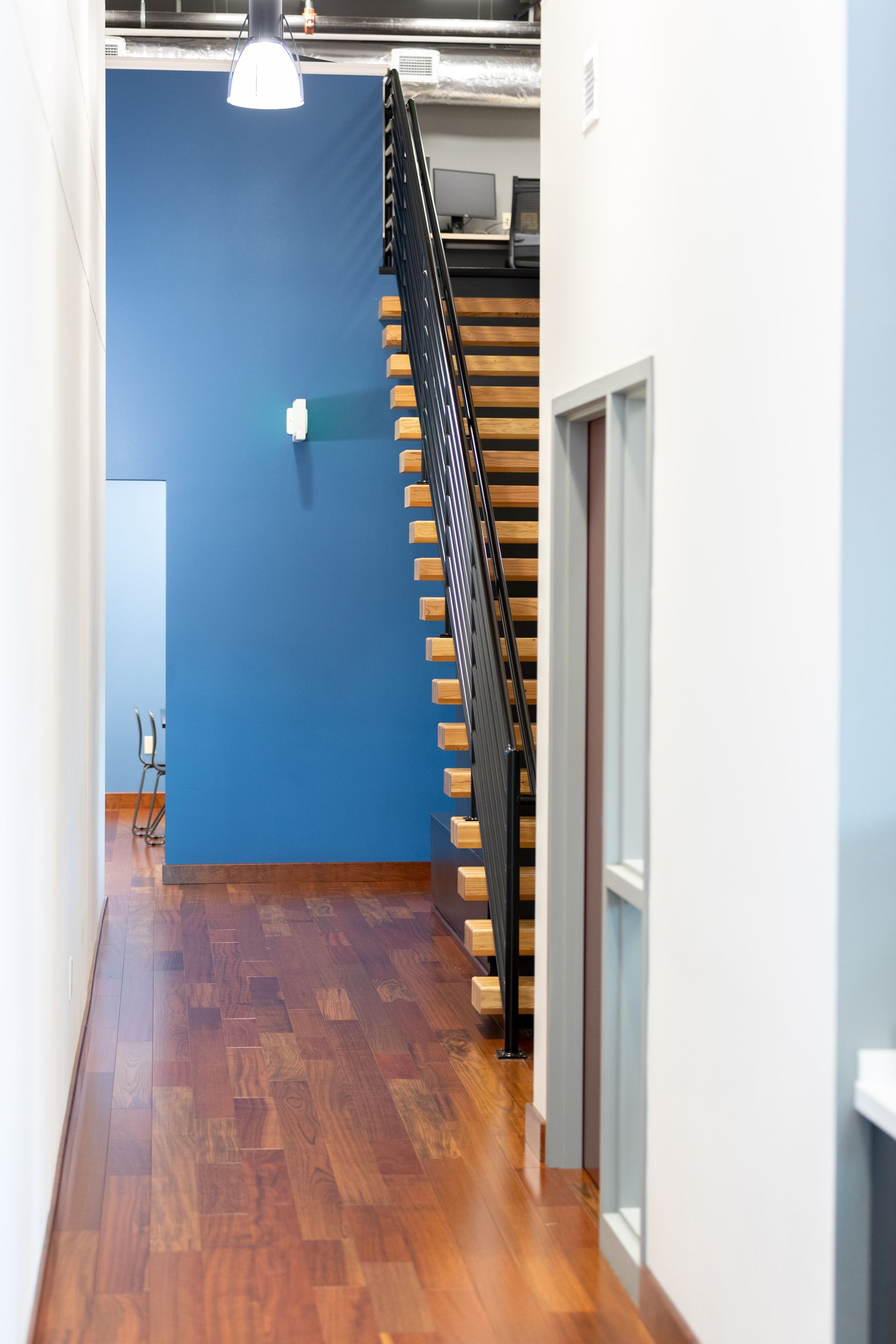 Office hallway with wooden floor, blue wall, and staircase with black railings.