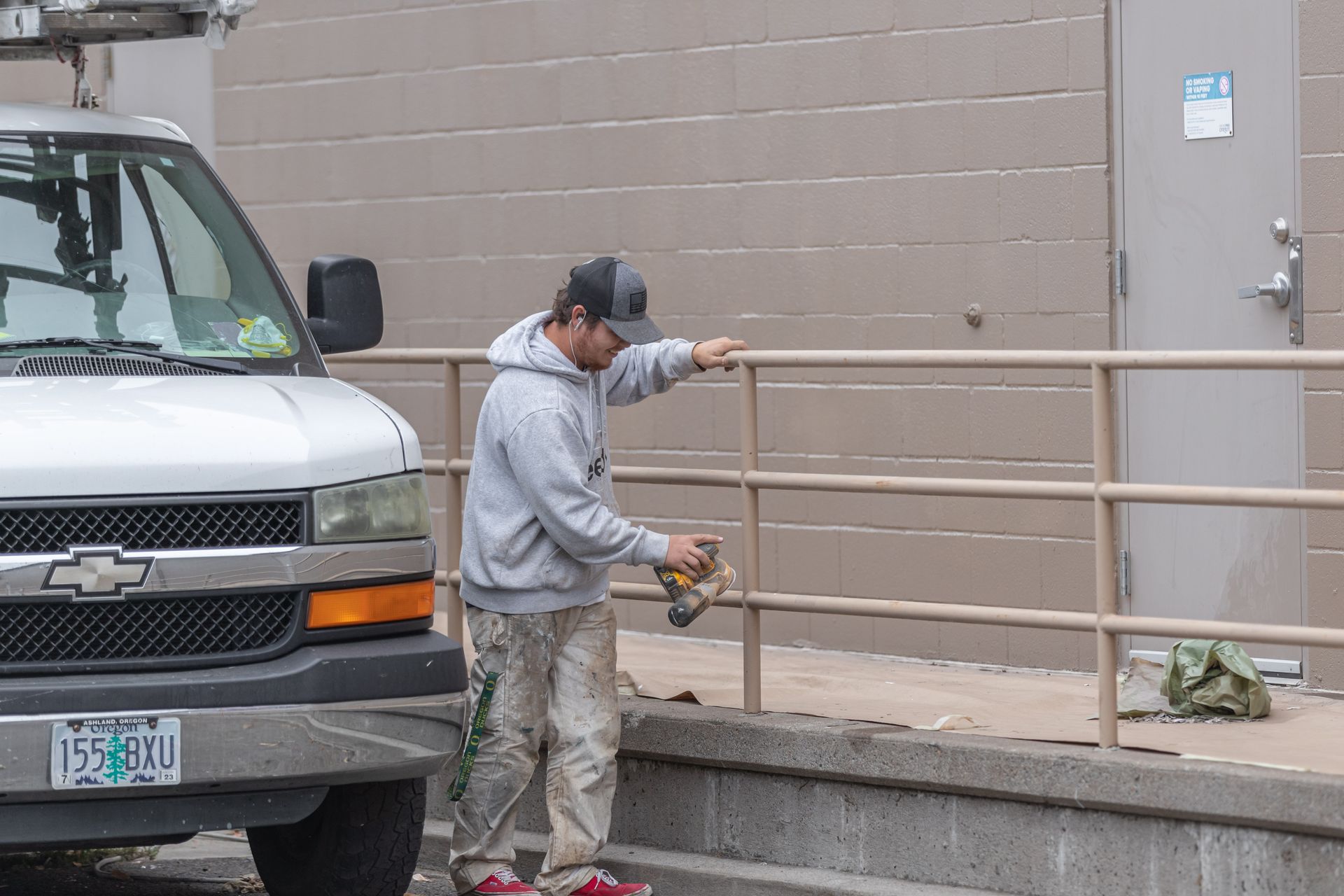 Worker in gray hoodie painting a metal railing beside a building and parked van.