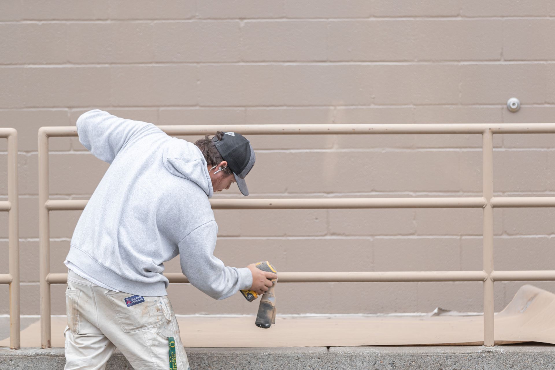 Person in a gray hoodie spray-painting a beige wall behind a metal railing.