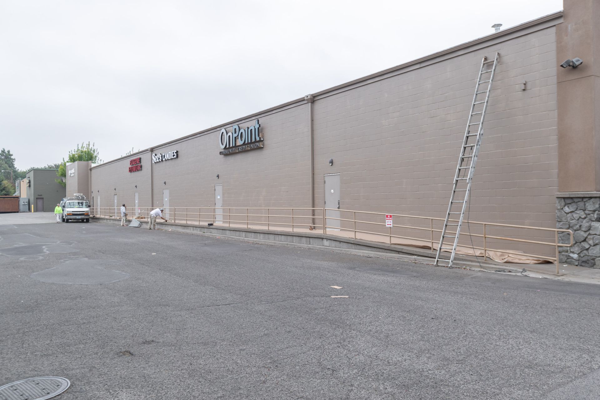 Large beige storefront wall with signs, parked cars, and a ladder leaning against the building.