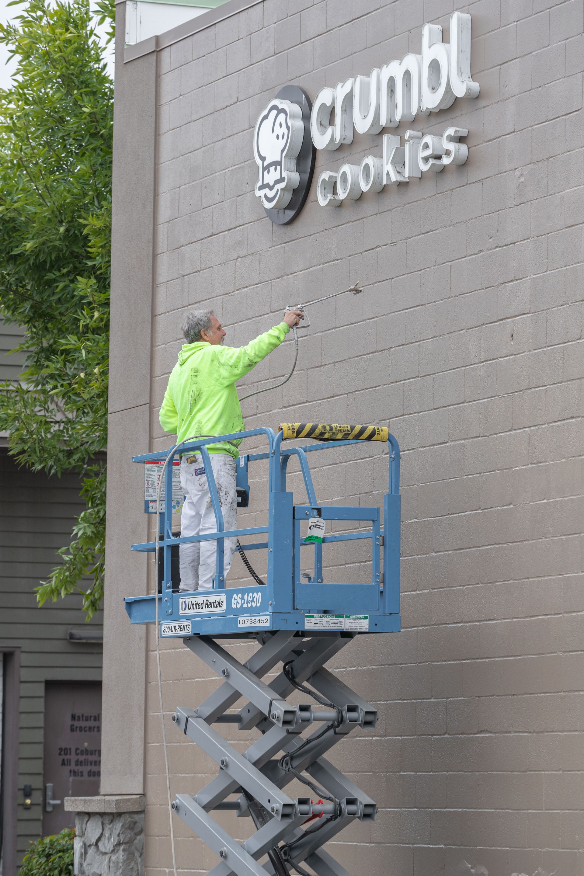 Worker in neon jacket installing signage on a Crumbl Cookies store wall from a lift.