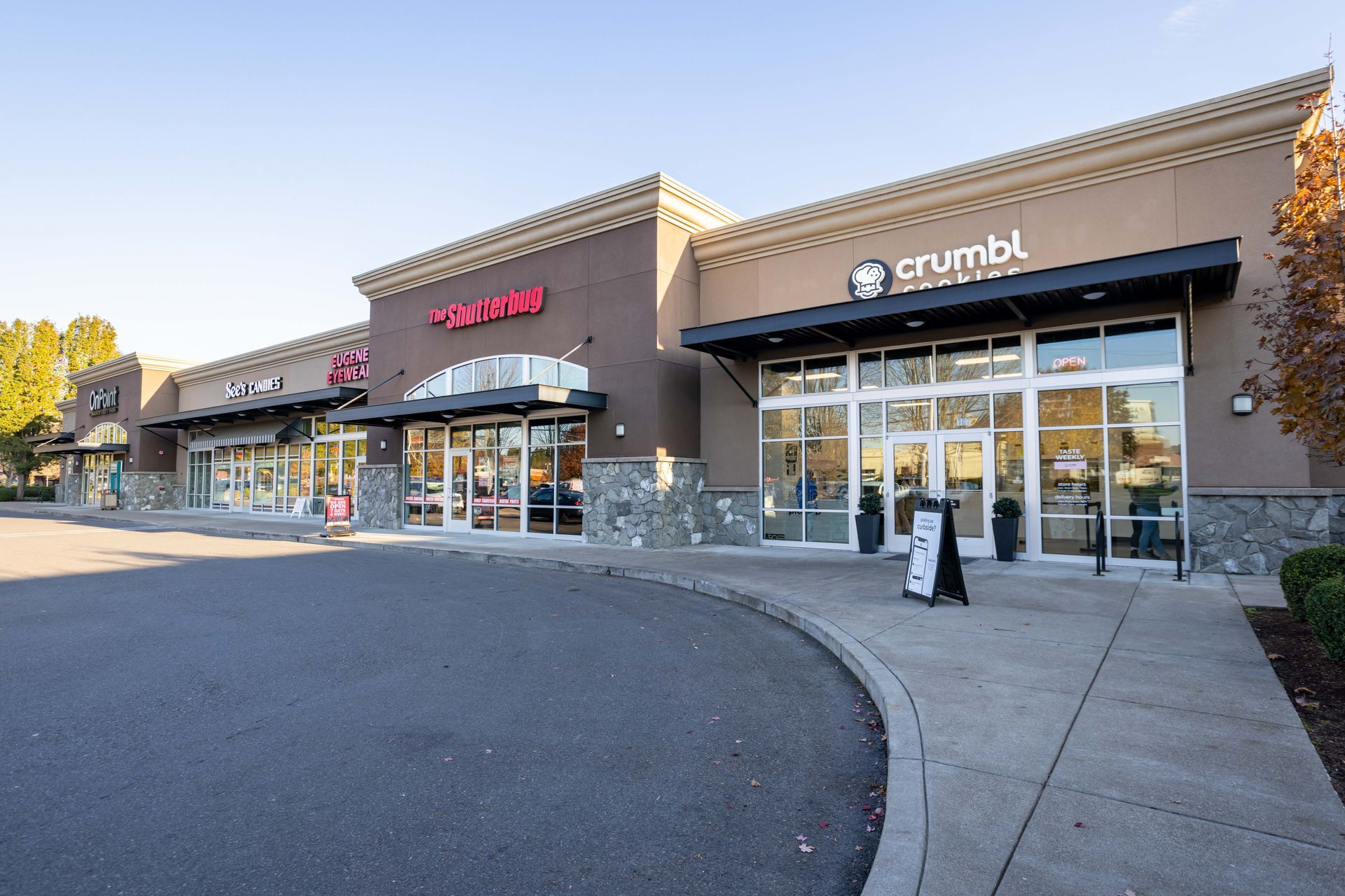 Strip mall storefronts, including Crumbl Cookies and a red sign, along a curved sidewalk in daylight