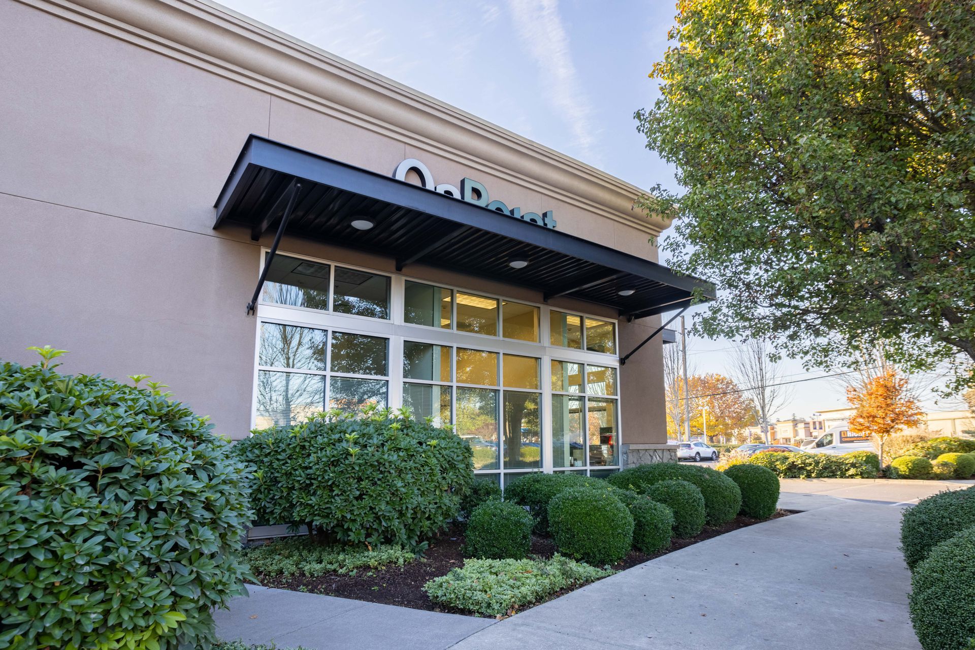 Office building entrance with black awning, glass doors, and trimmed bushes along a walkway