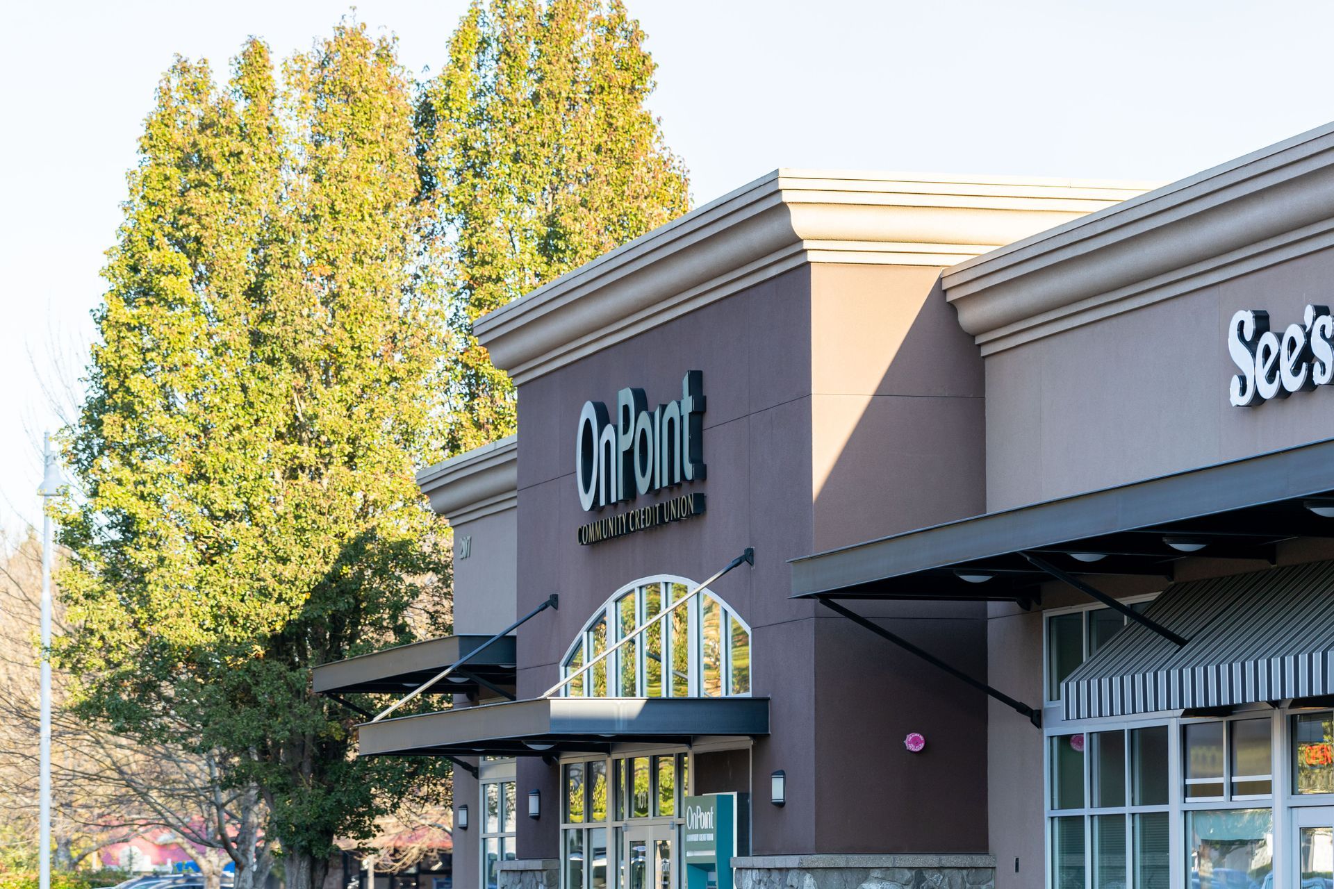 Shopping center storefronts with teal signage and a striped awning beside tall green trees