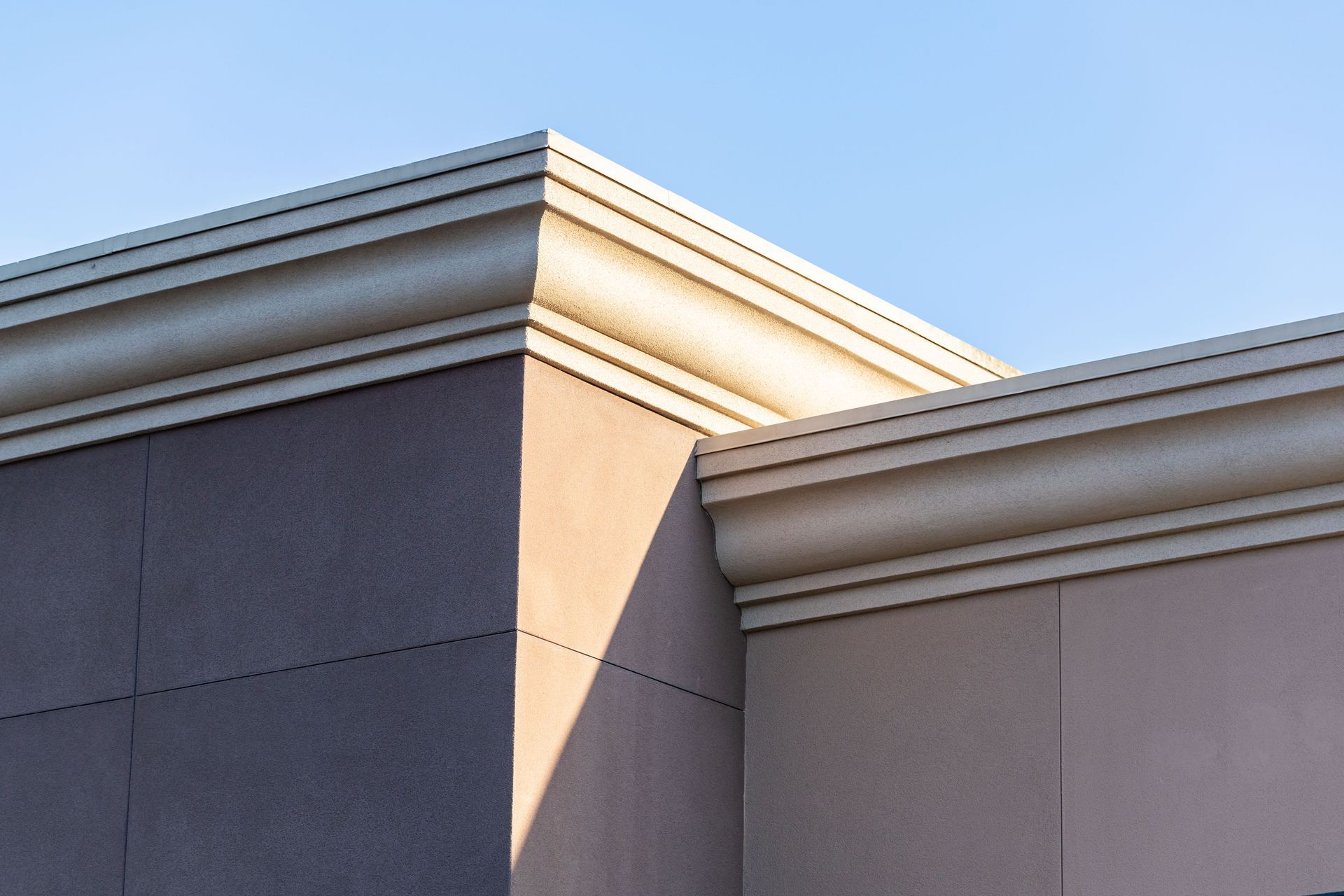 Close-up of a beige building roofline and cornice against a blue sky, with sunlight casting a shadow.