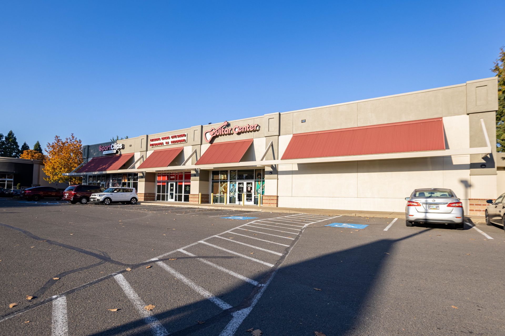 Strip mall storefronts with red awnings and parked cars in a sunlit lot