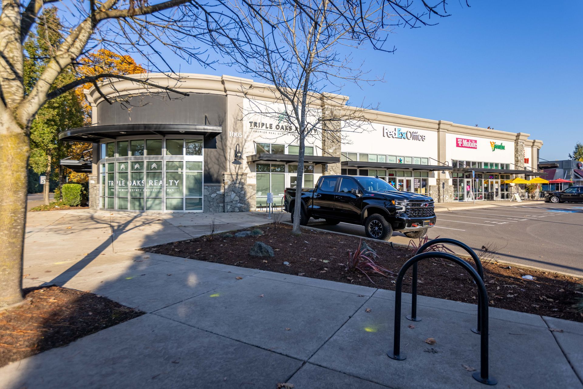 Outdoor shopping center with storefronts, parked SUV, and bike rack on a sunny day