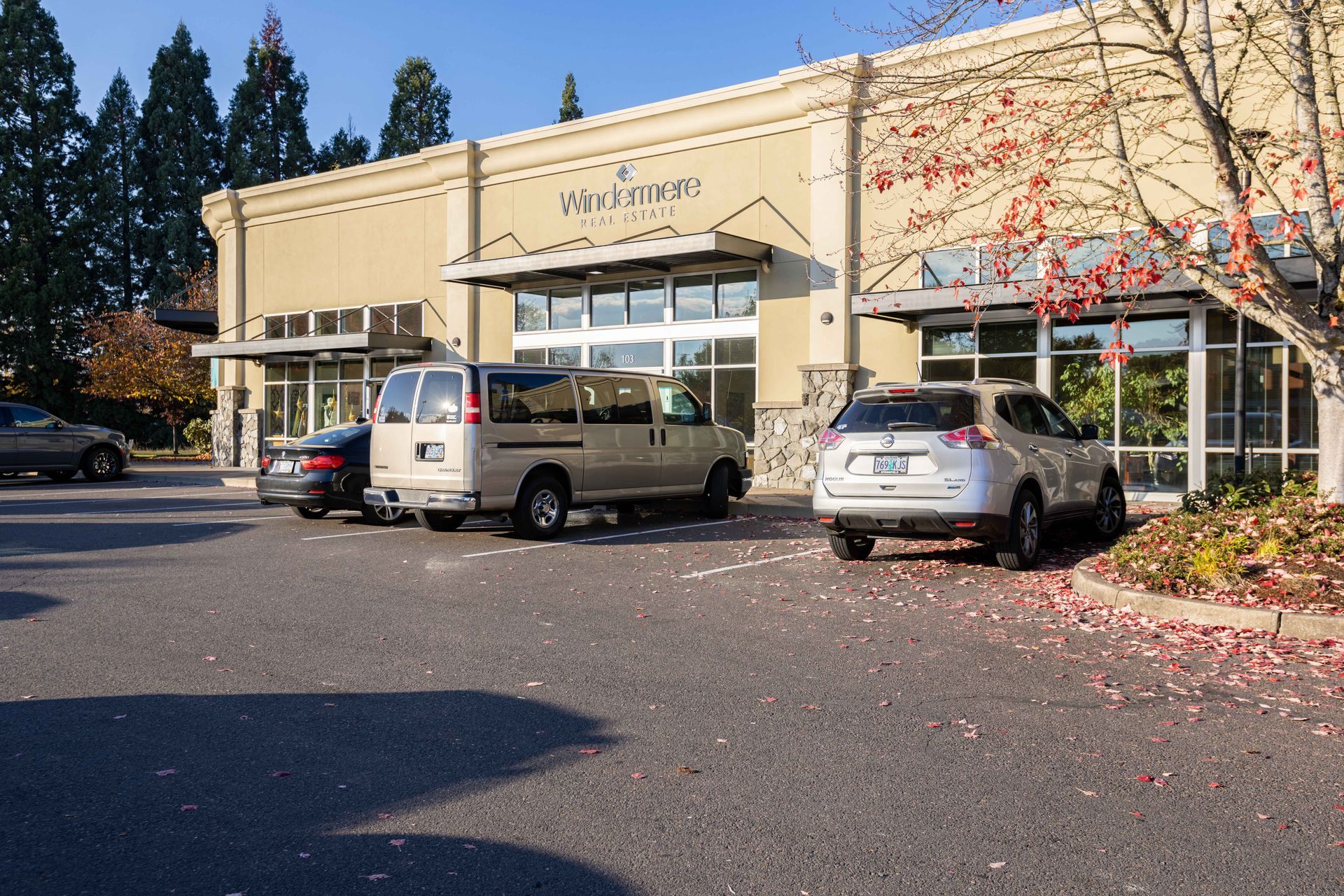 Parking lot outside a beige commercial building with parked cars and autumn trees in the background
