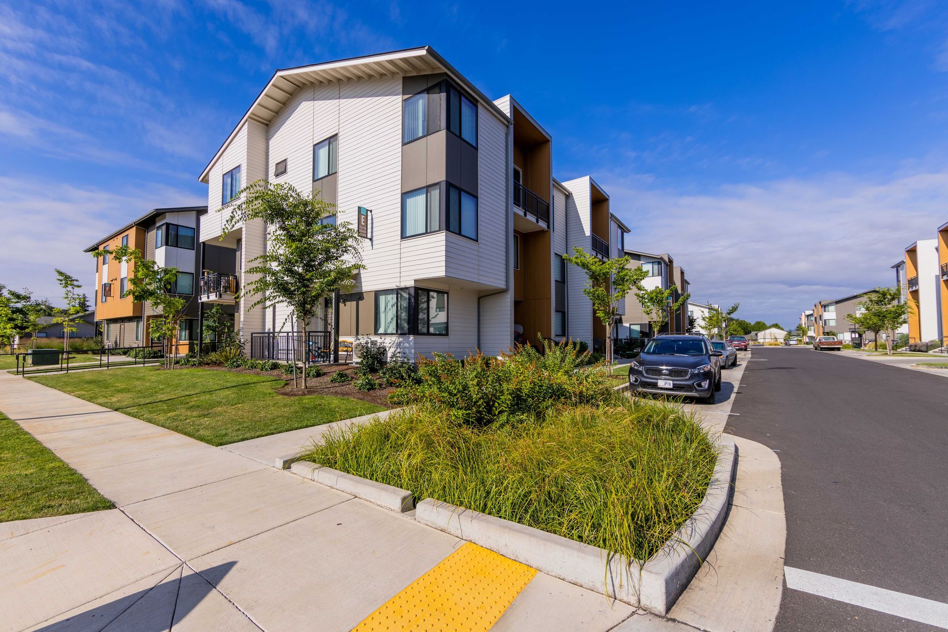Modern apartment buildings on a sunny street corner with sidewalks, landscaping, and parked cars