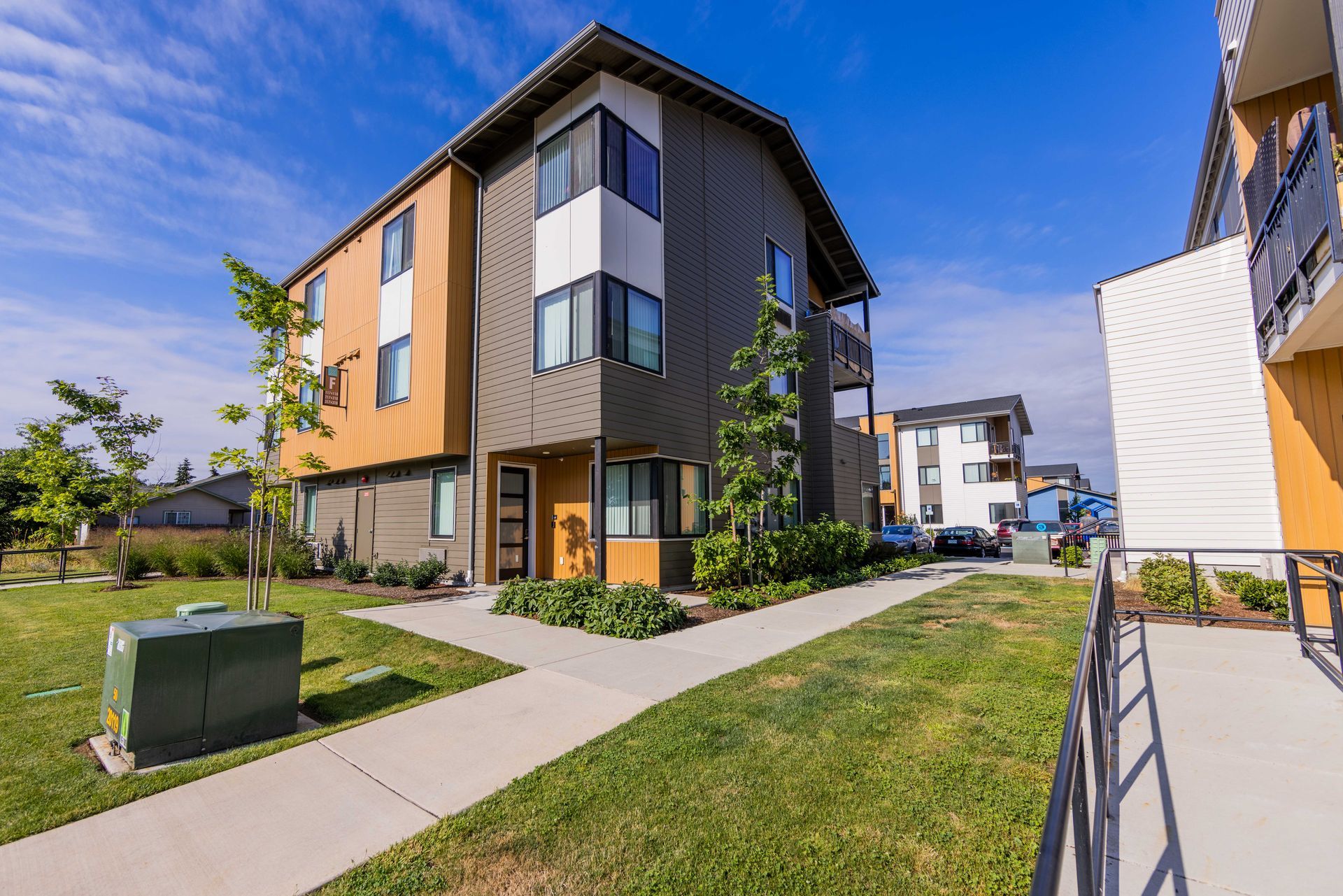 Modern apartment building with tan and gray siding, grassy walkway, and blue sky on a sunny day
