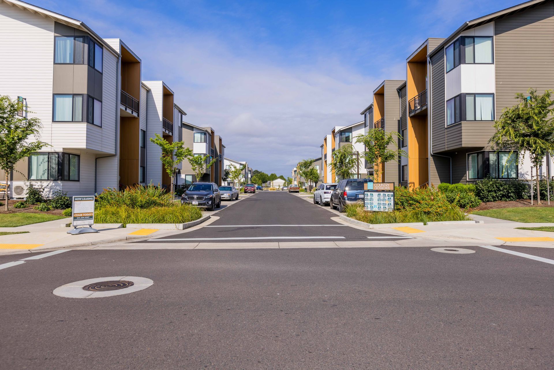 Modern apartment complex with a wide street, parked cars, and palm trees under a blue sky