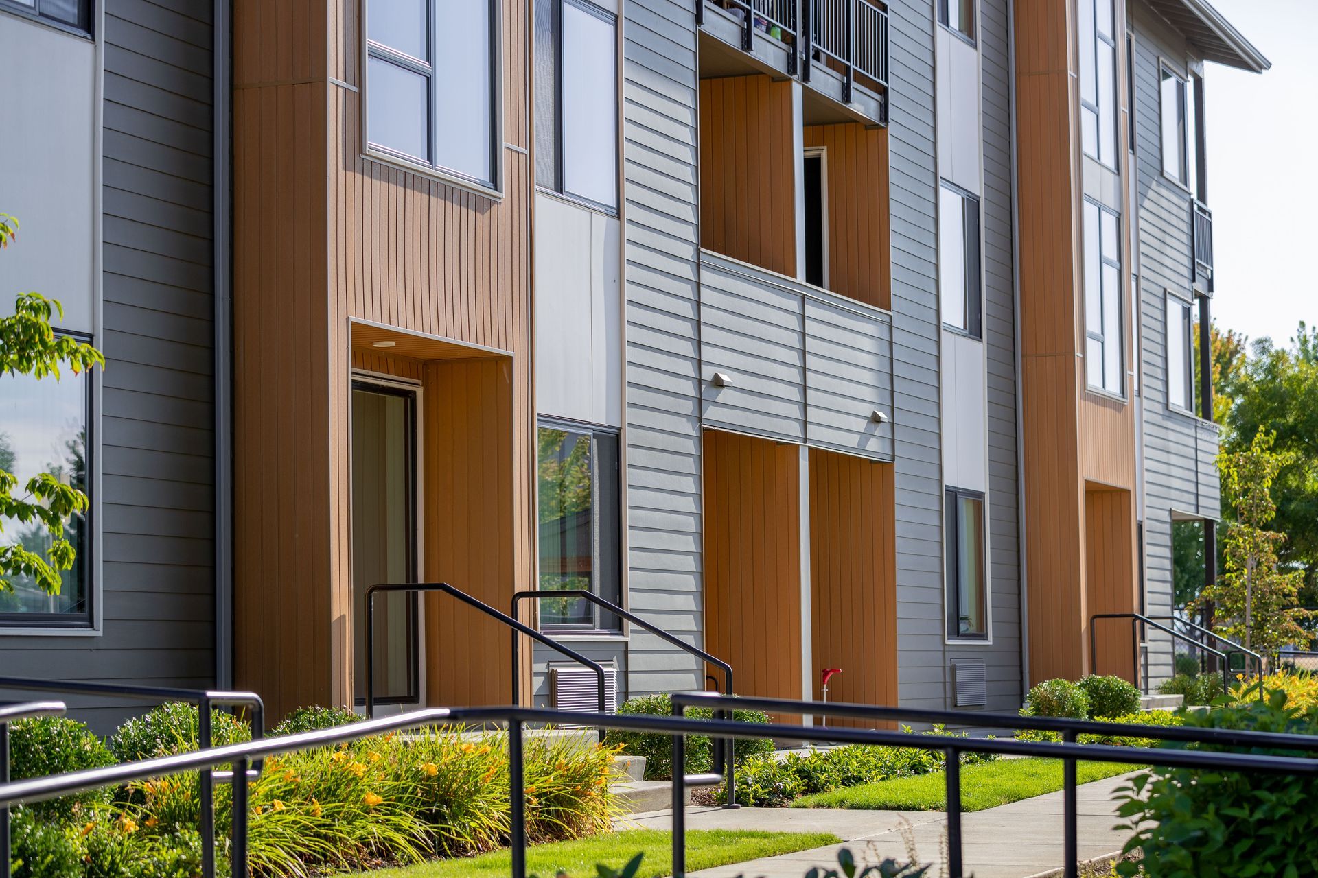 Modern apartment building with gray and wood siding, balconies, and landscaped shrubs out front