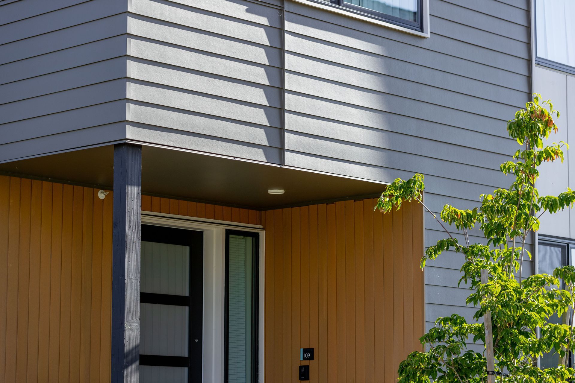 Modern building entrance with gray siding, wood paneling, and a small tree nearby