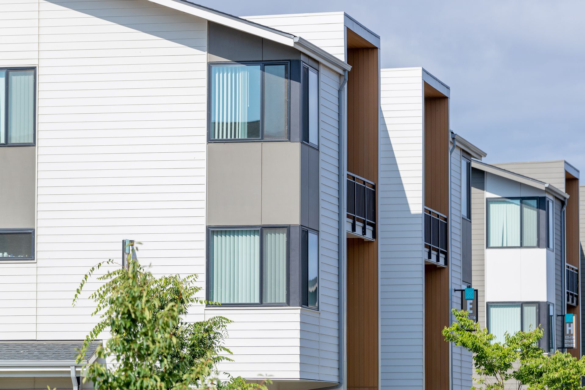 Modern apartment buildings with white and gray siding and balconies, framed by green trees