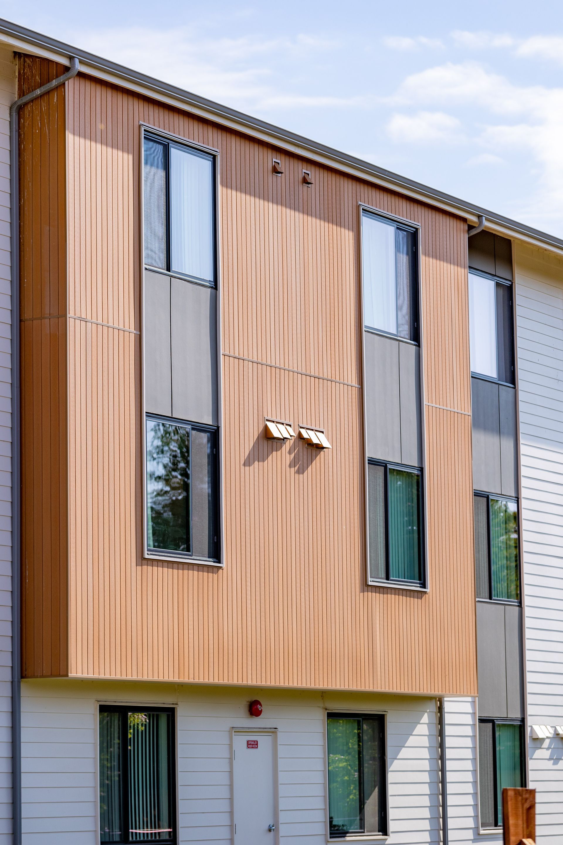 Modern apartment building facade with wood and gray panels, narrow windows, and a clear sky above