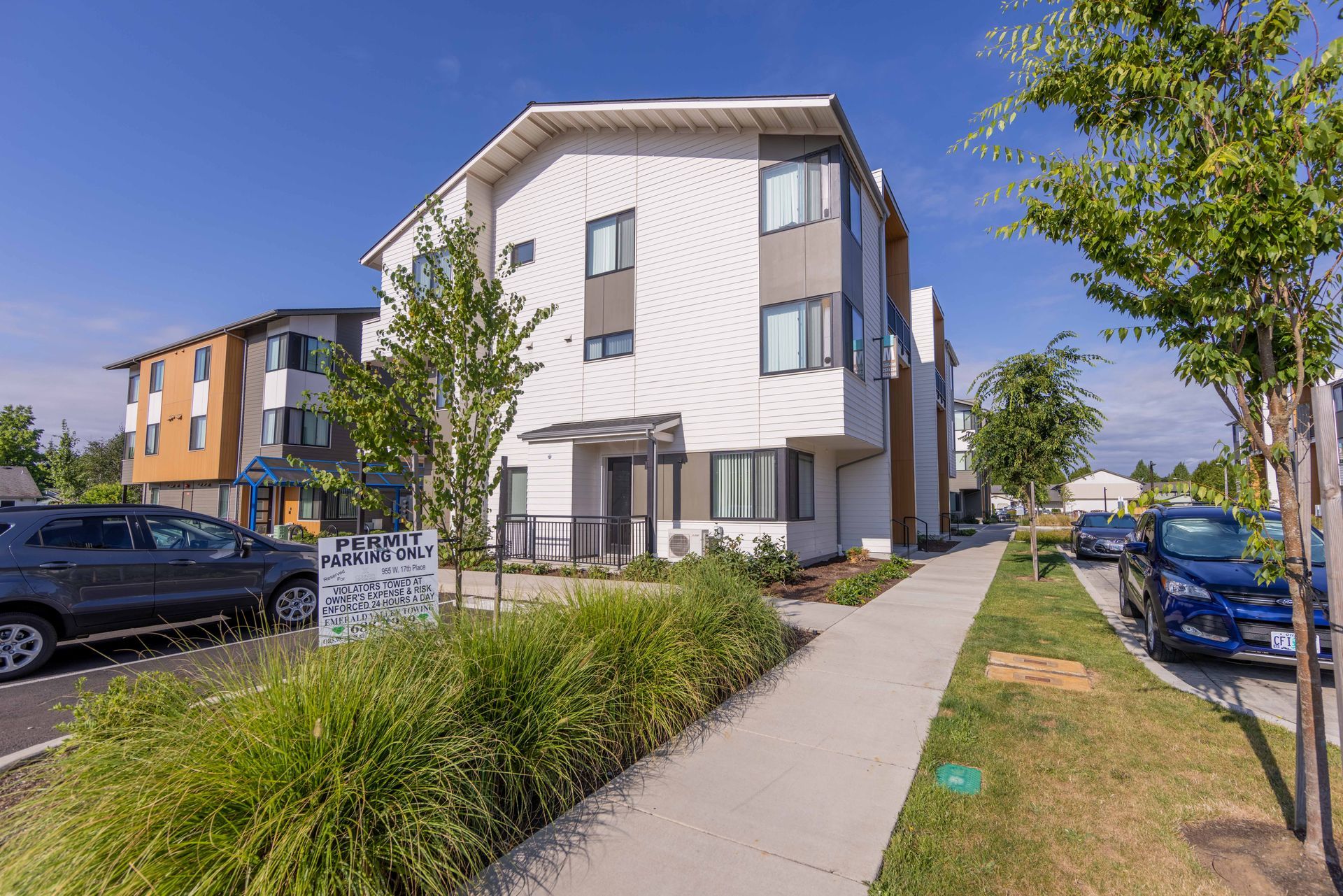 Modern apartment buildings beside a sidewalk with parked cars and trees on a sunny day