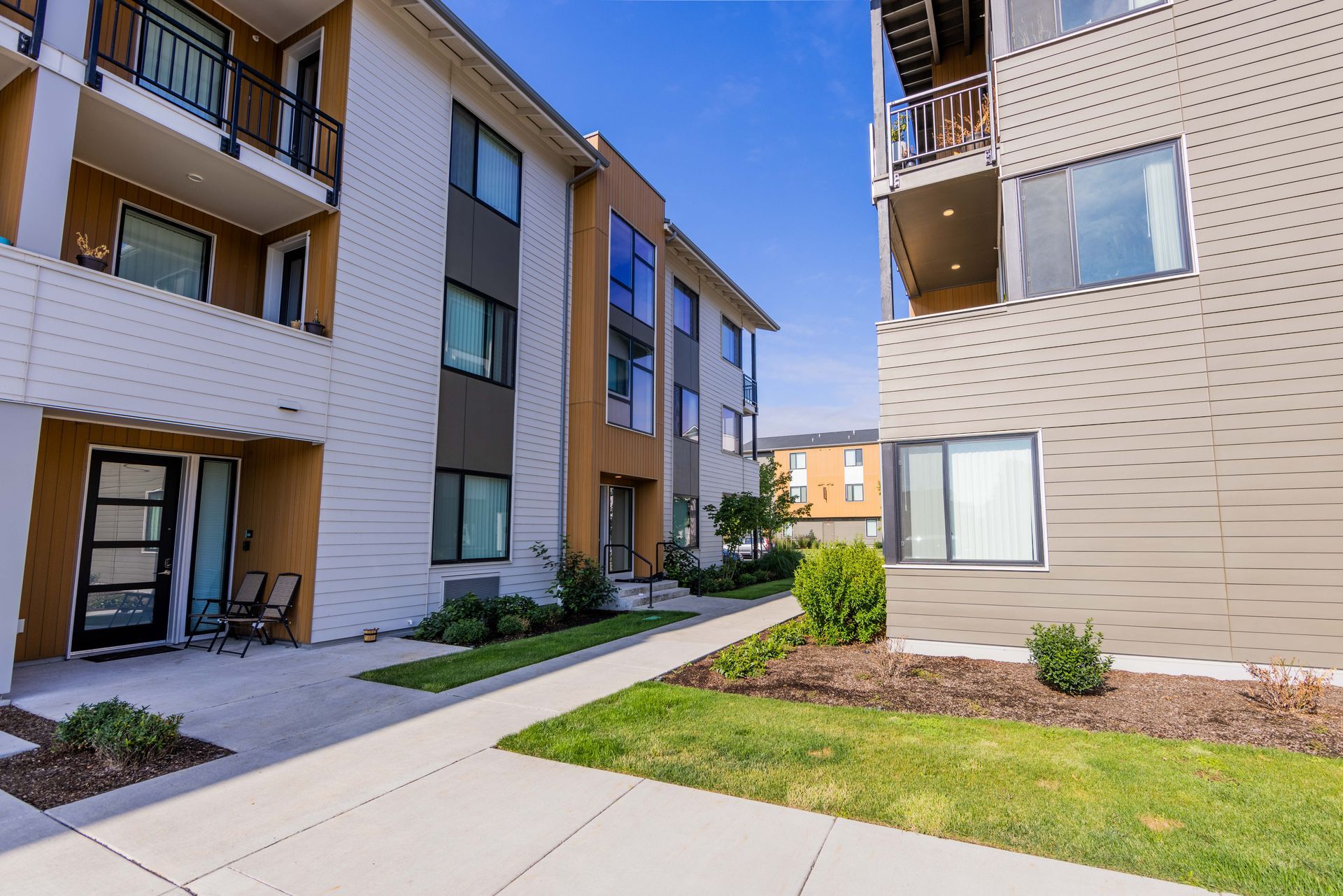 Apartment complex courtyard with tan brick buildings, walkway, and green lawn under a blue sky