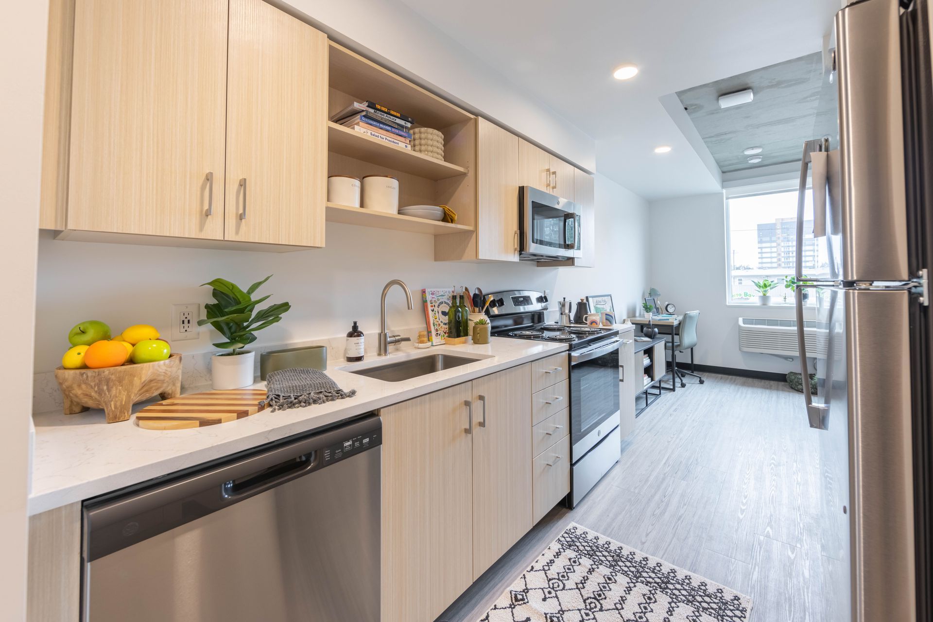 Modern galley kitchen with light wood cabinets, stainless appliances, and a bright dining nook at the end.