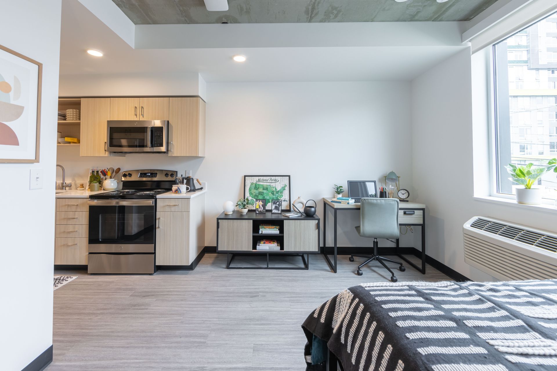Modern studio apartment with kitchen, desk, chair, bed, and large window.