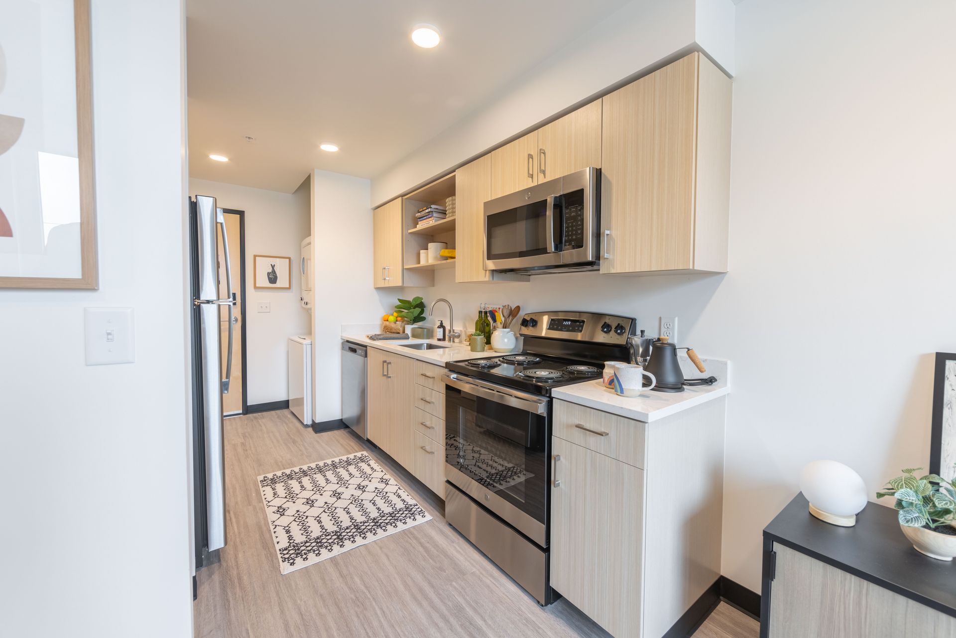 Narrow modern kitchen with light wood cabinets, stainless appliances, and a patterned runner rug.