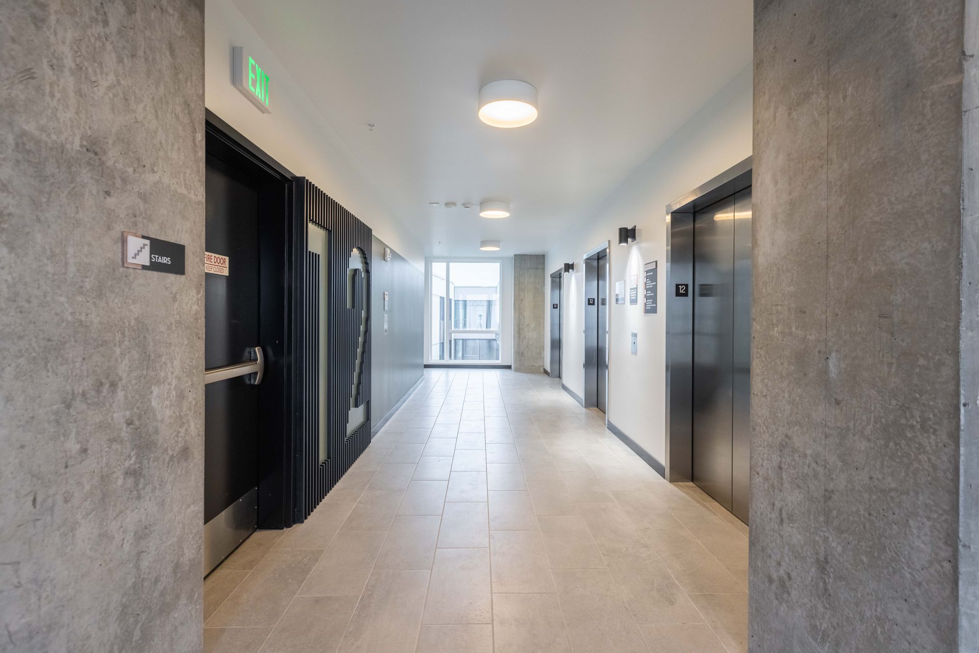 Modern apartment hallway with elevators, black doors, and a bright window at the end