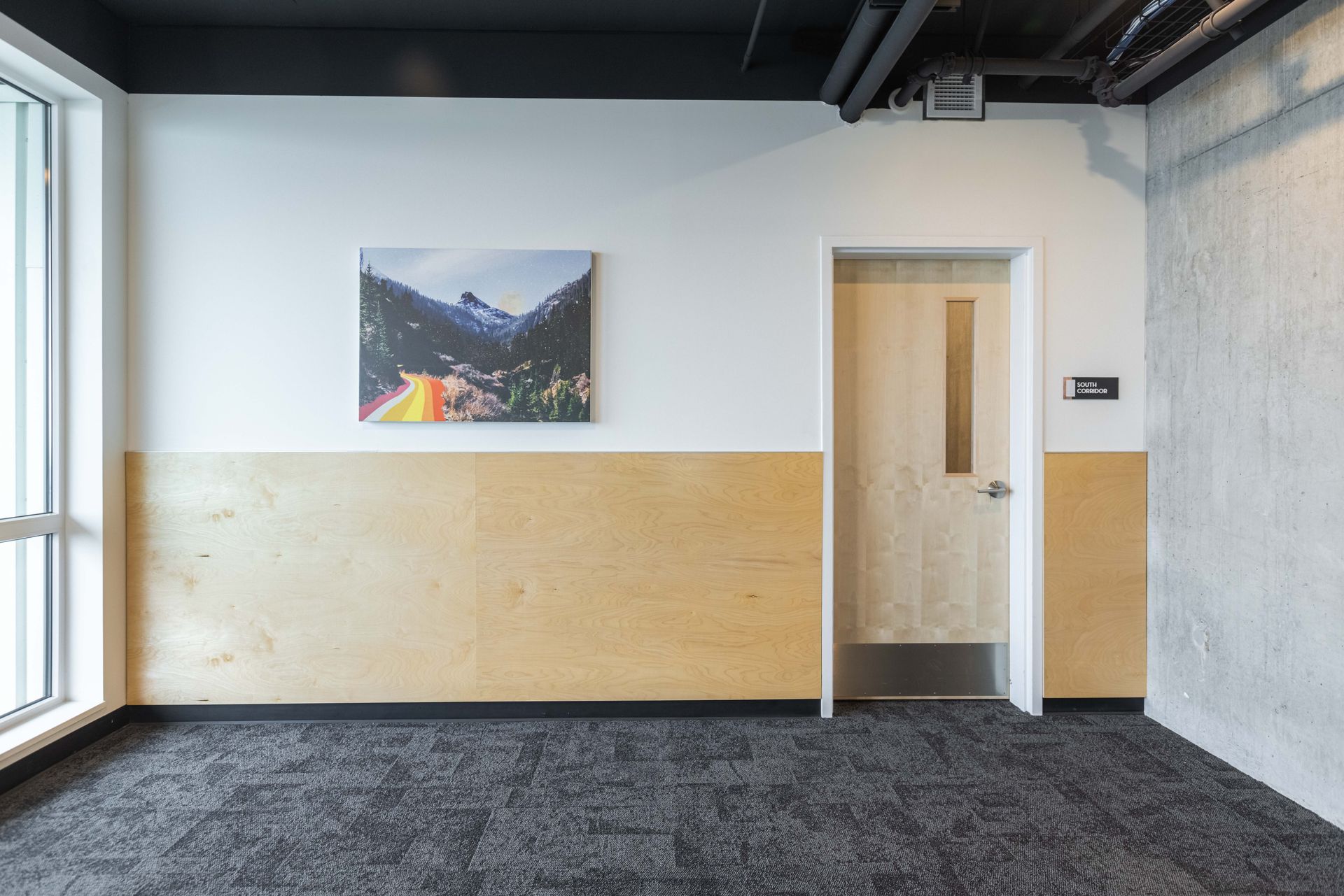 Empty office hallway with a wood-paneled wall, framed landscape photo, and door beside a window.