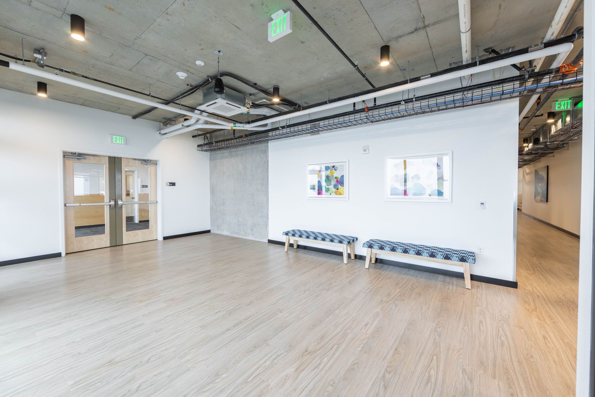 Bright open office or studio with light wood floors, white walls, exposed ceiling, and benches along the wall