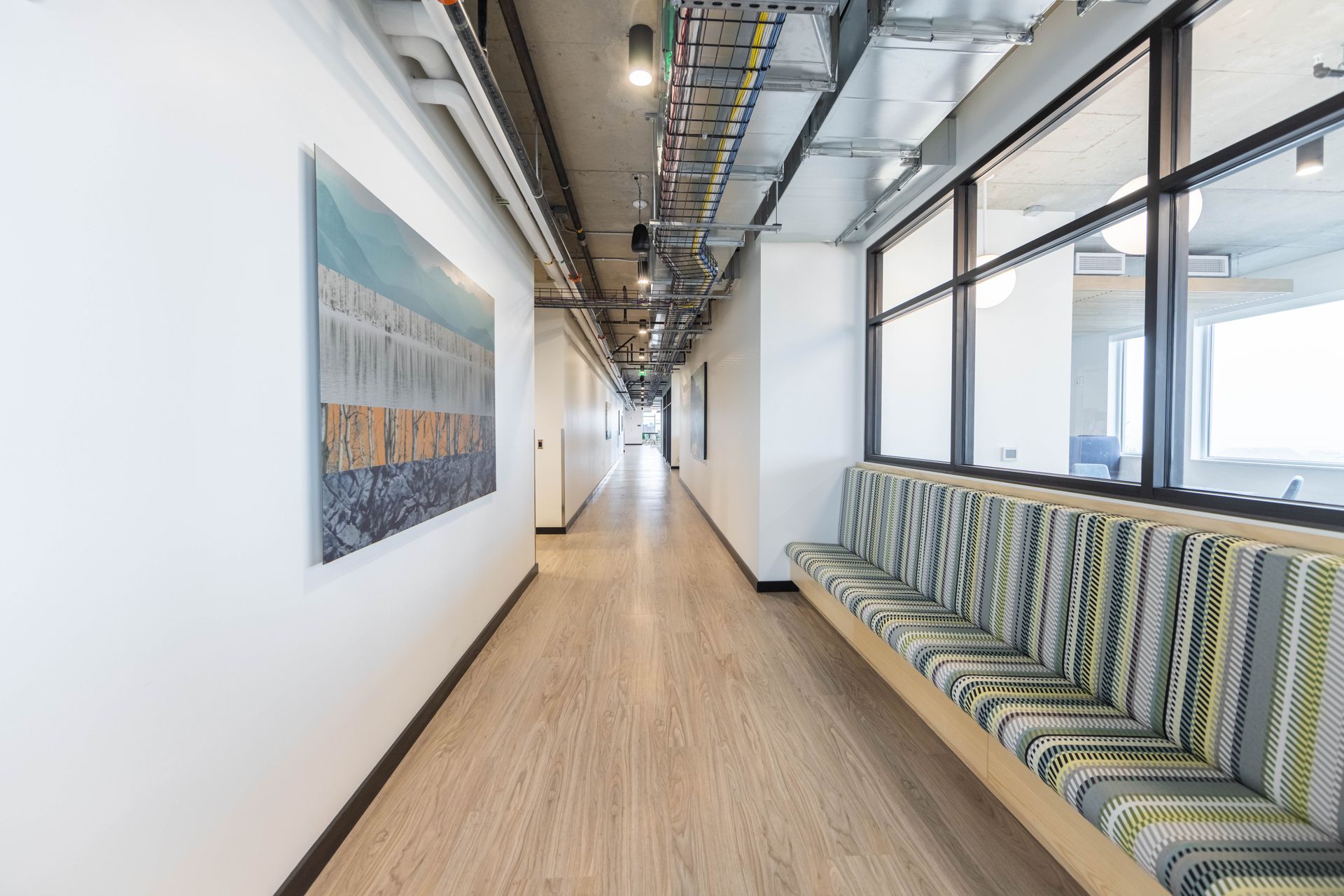 Modern office hallway with striped bench seating, wall art, and windows on the right