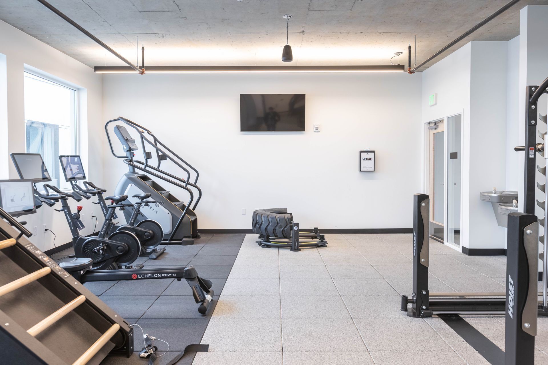 Empty gym room with exercise machines, a wall-mounted TV, and a rubber floor mat