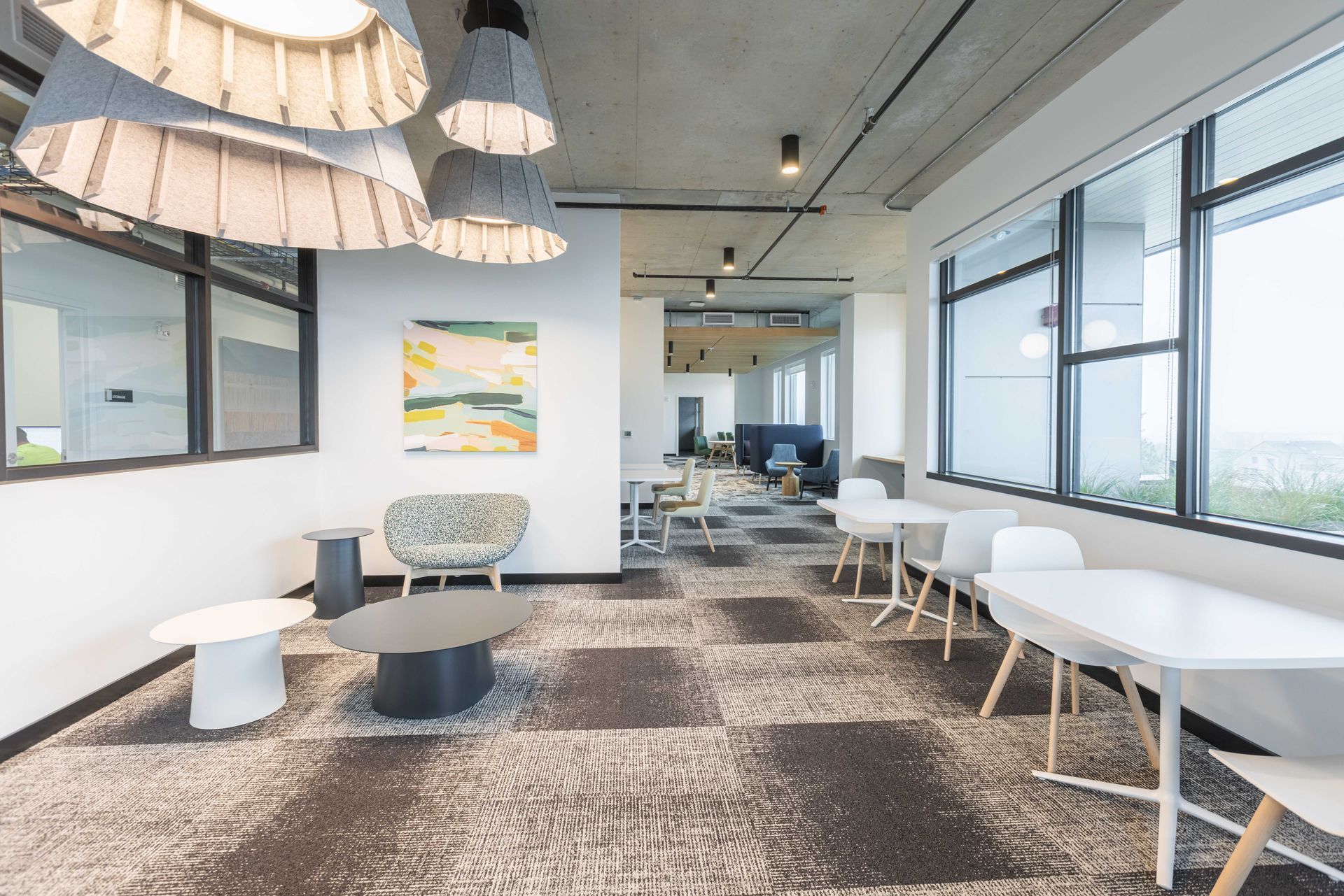 Modern office lounge with white tables, gray chairs, patterned rug, and large windows overlooking a bright city view