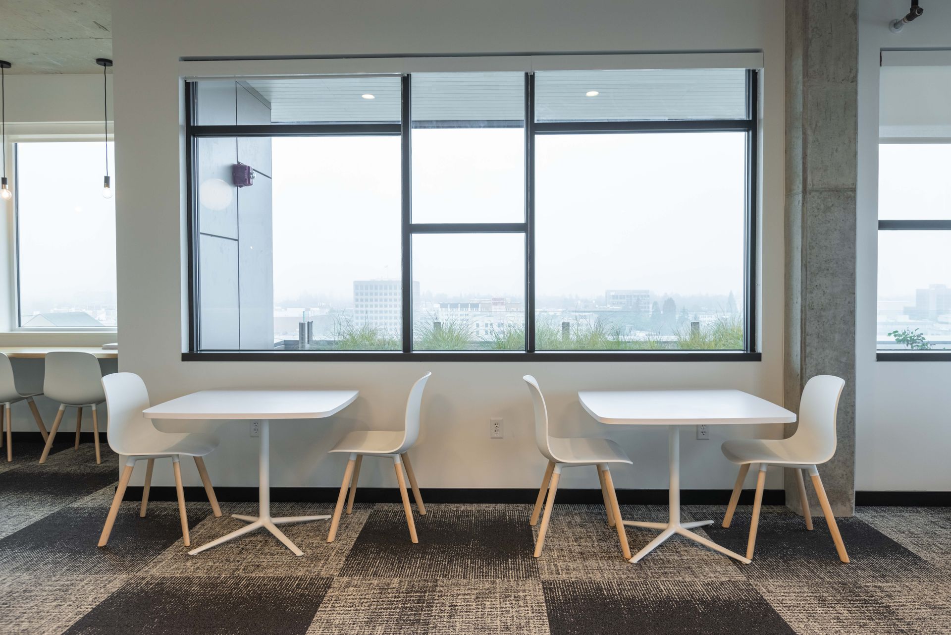 Modern office lounge with white tables and chairs beside large windows overlooking a city view