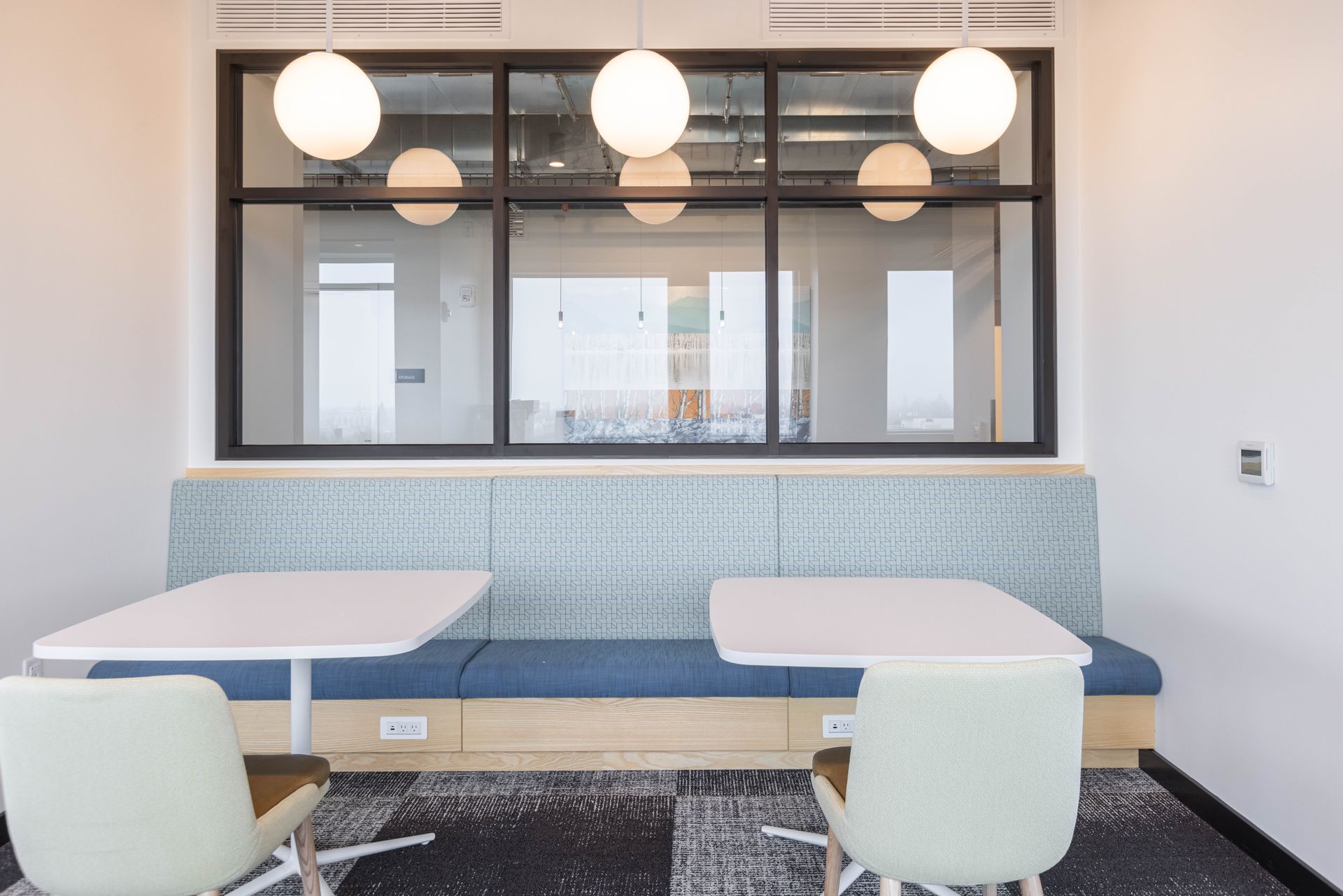 Modern office lounge with two tables, cushioned bench seating, chairs, and hanging globe lights