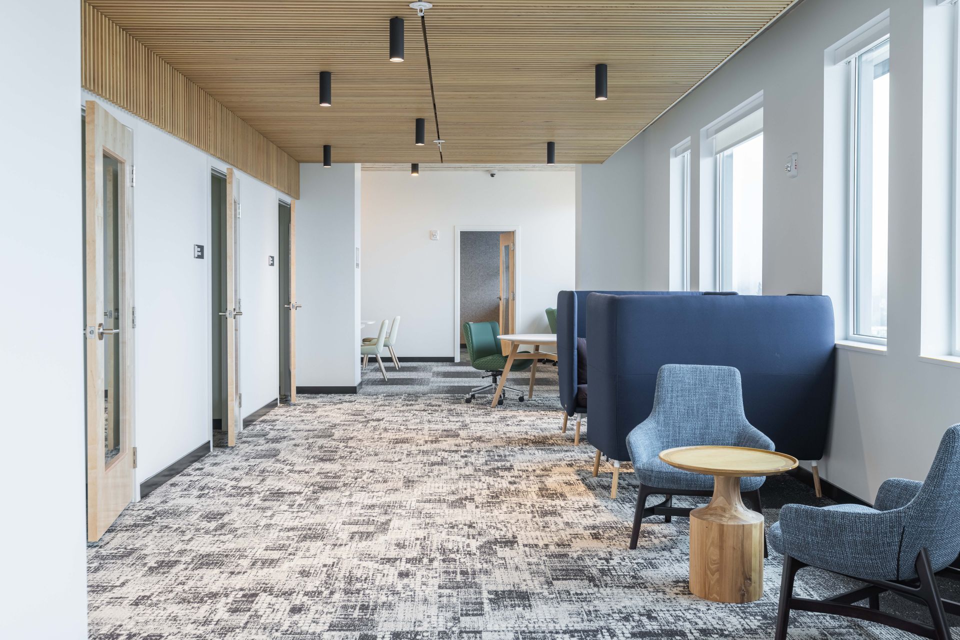Modern office hallway with white walls, wood ceiling, patterned carpet, chairs, and blue booth seating