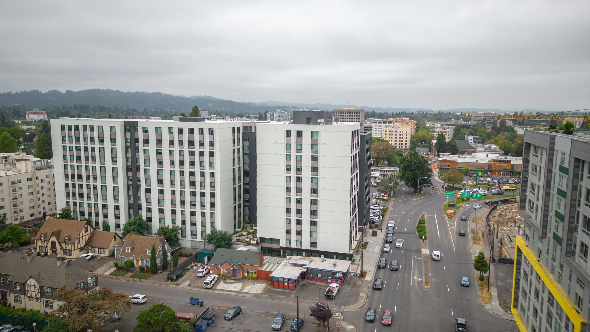 Aerial view of a city street with tall apartment buildings, cars, and a cloudy sky.