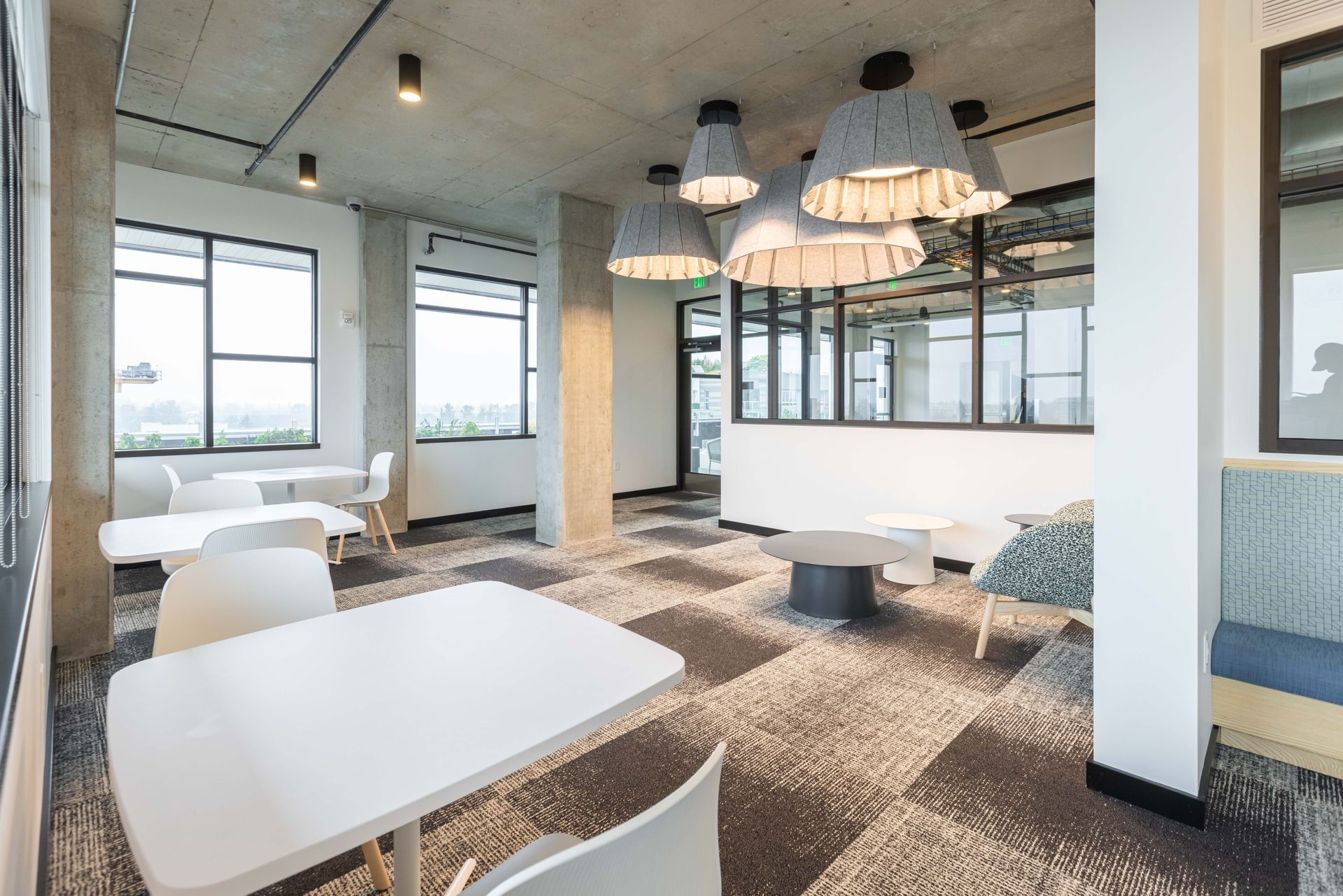 Modern lounge with white tables, pendant lights, patterned carpet, and large windows overlooking a city view