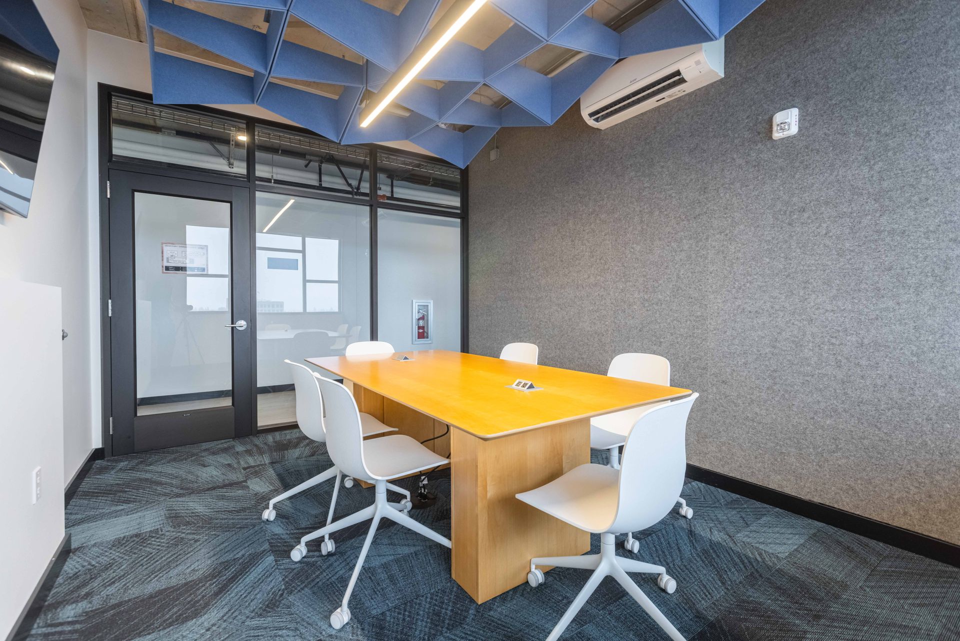 Modern conference room with a wooden table, white chairs, glass partition, and blue ceiling panels