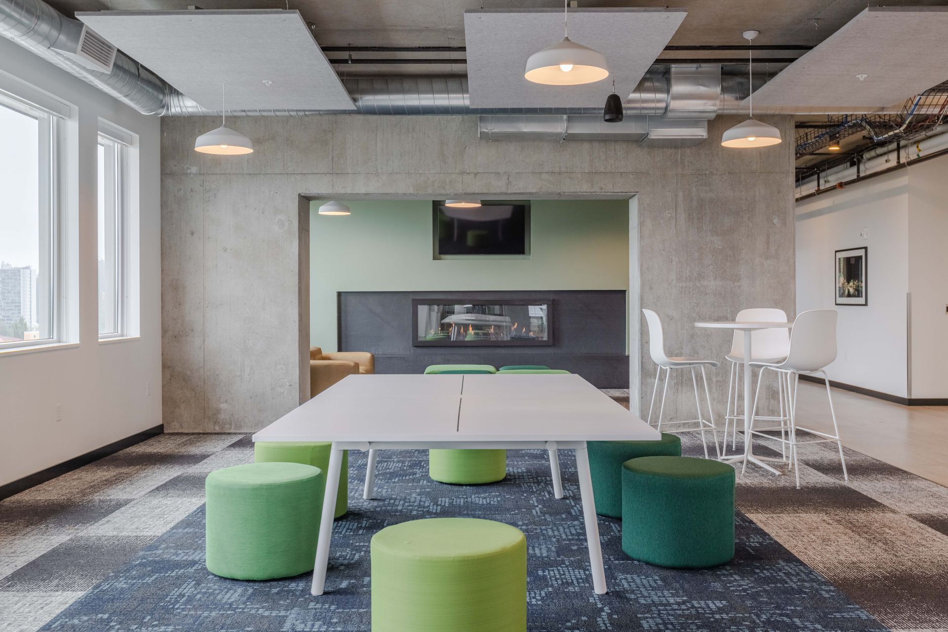 Modern office lounge with white table, green stools, wall-mounted TV, and white chairs near windows