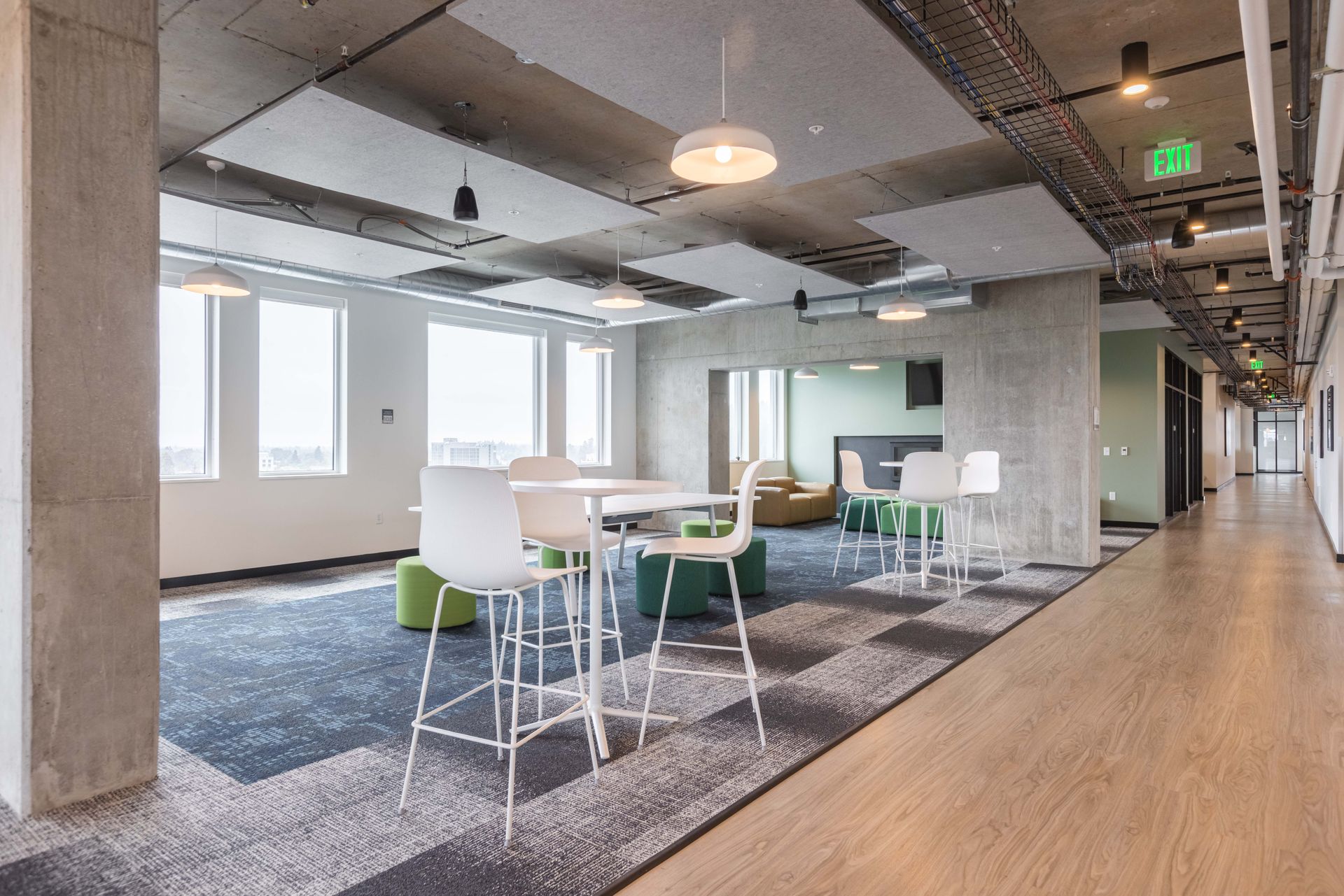 Modern office lounge with white chairs, green tables, carpet, and large windows under exposed ceiling beams