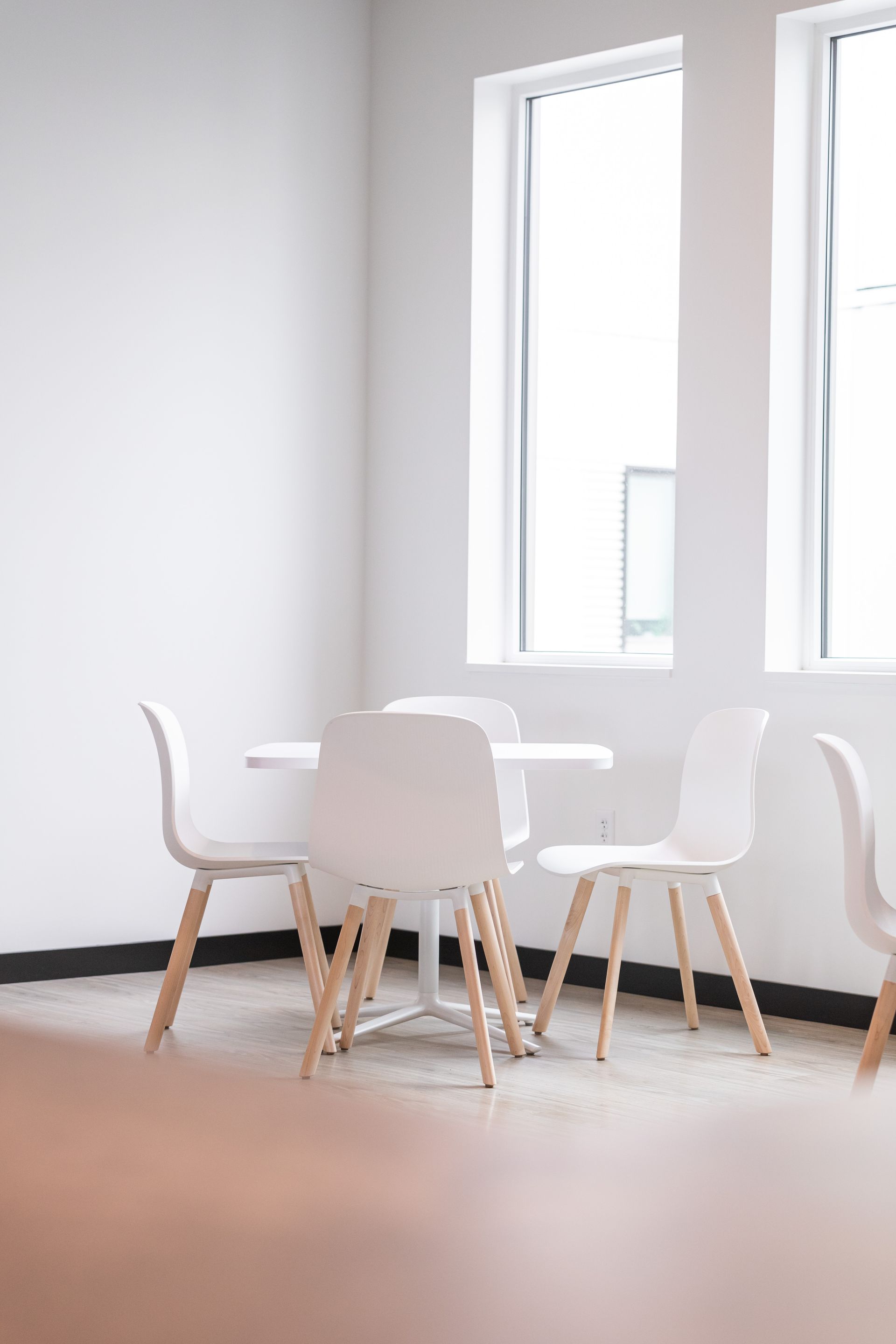 Minimal white dining area with a small round table and white chairs by tall windows.