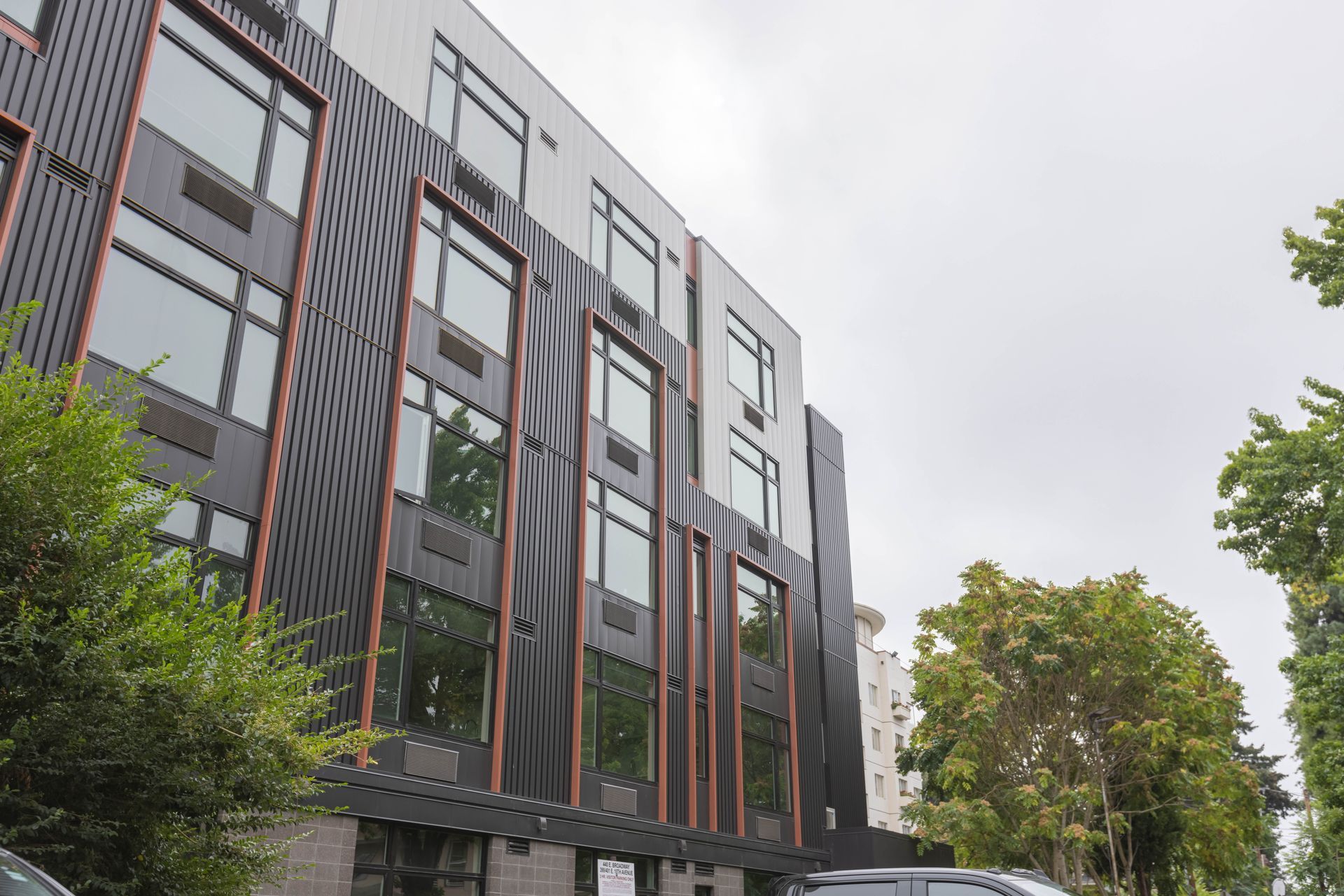 Modern apartment building with gray and brown facade, large windows, and trees along the street