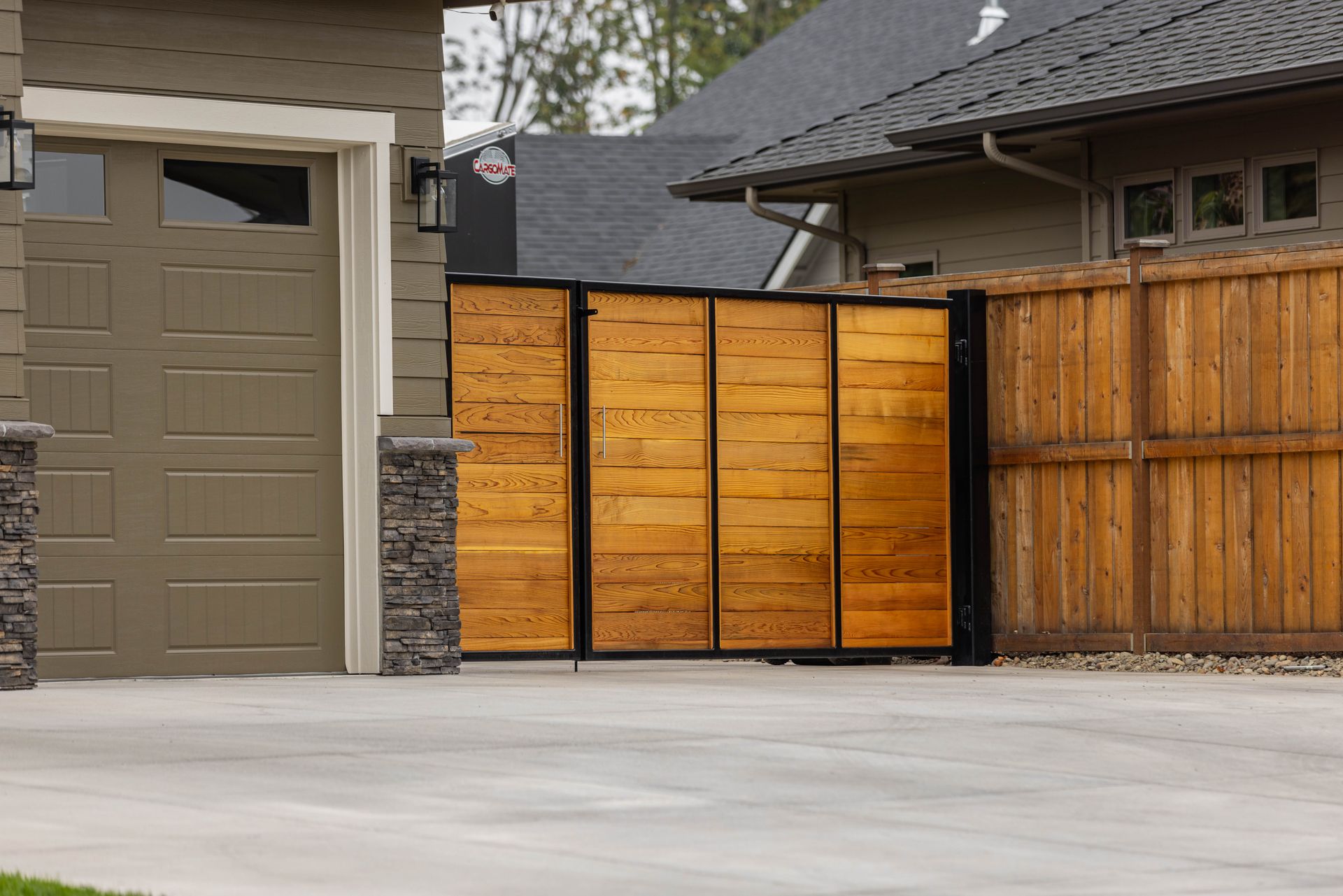 Residential driveway with a tan garage door and a wooden sliding gate beside a stone fence.