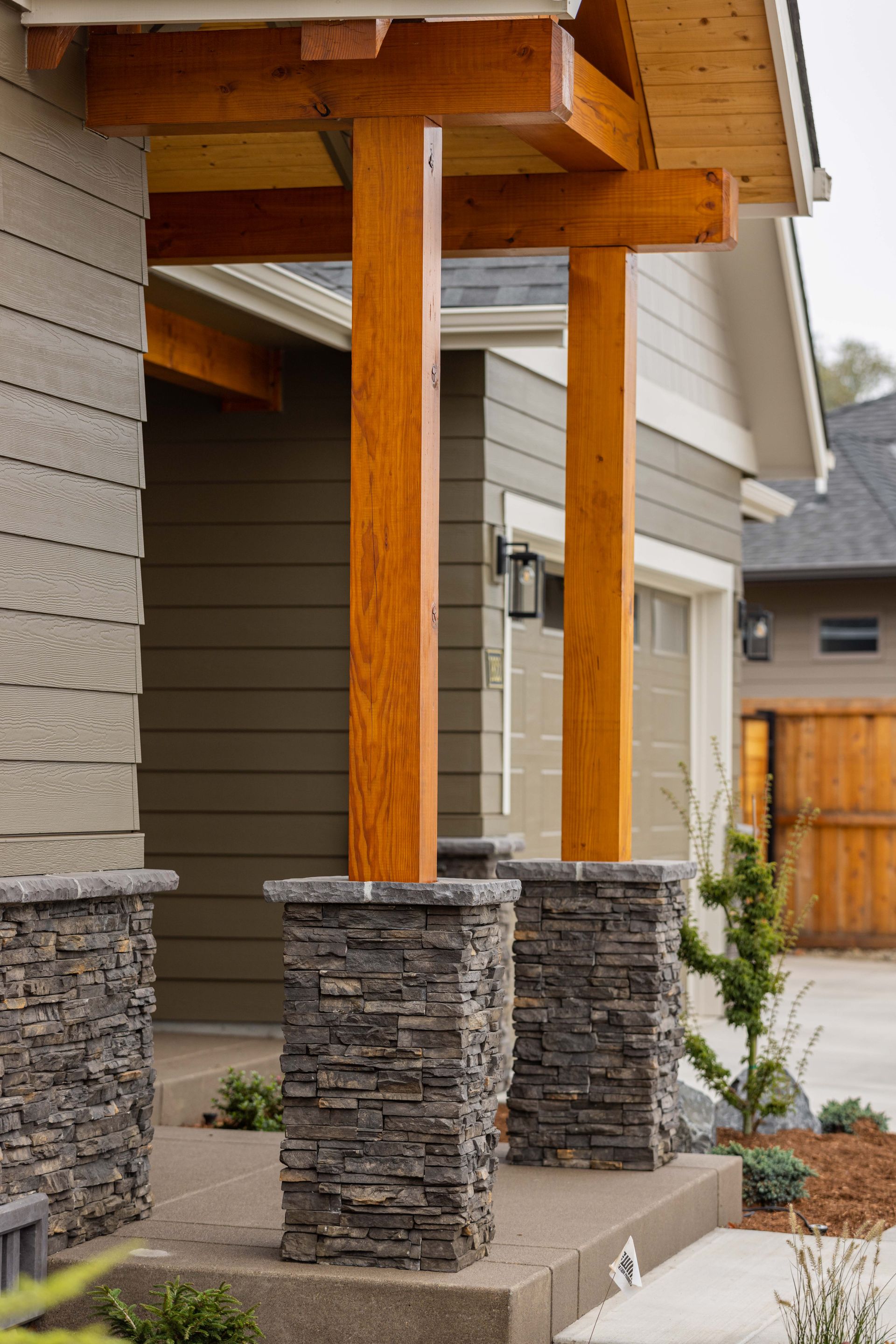 Stone-clad porch columns supporting a wooden overhang beside a beige house and driveway