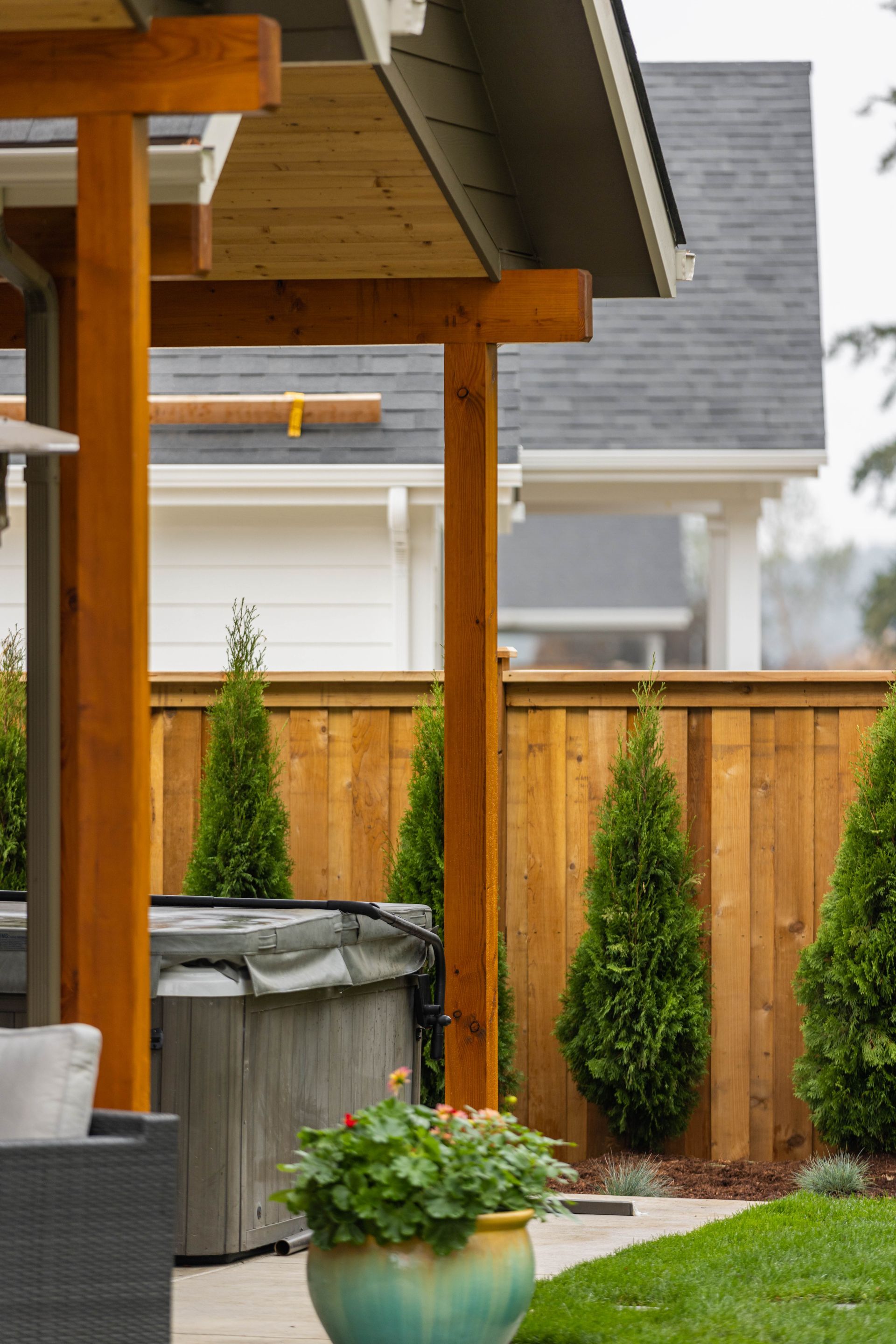 Covered backyard patio with wooden posts, grill, potted plants, and a privacy fence beside a house.