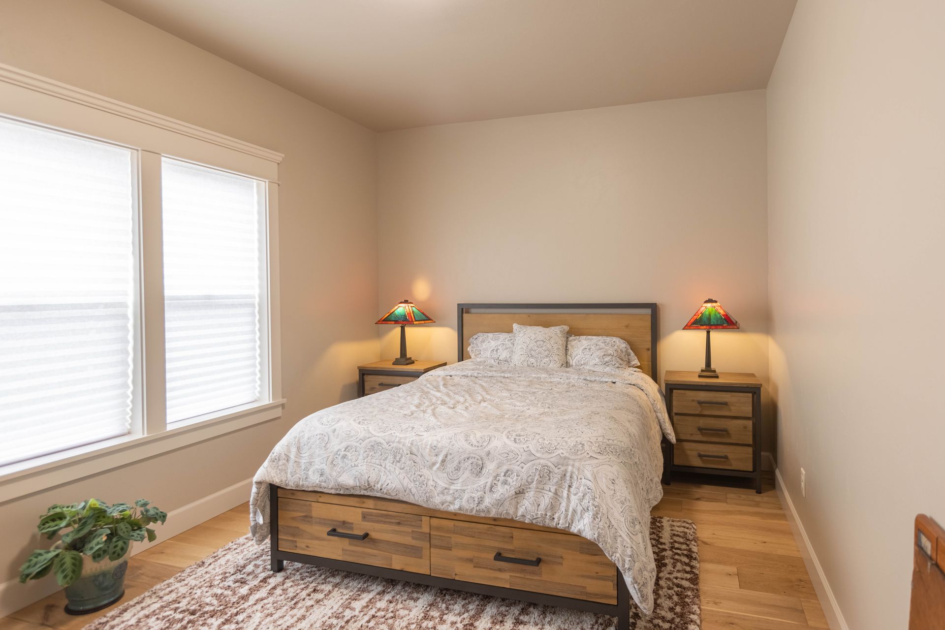 Bright bedroom with a wooden bed, bedside lamps, window blinds, and a potted plant on the floor